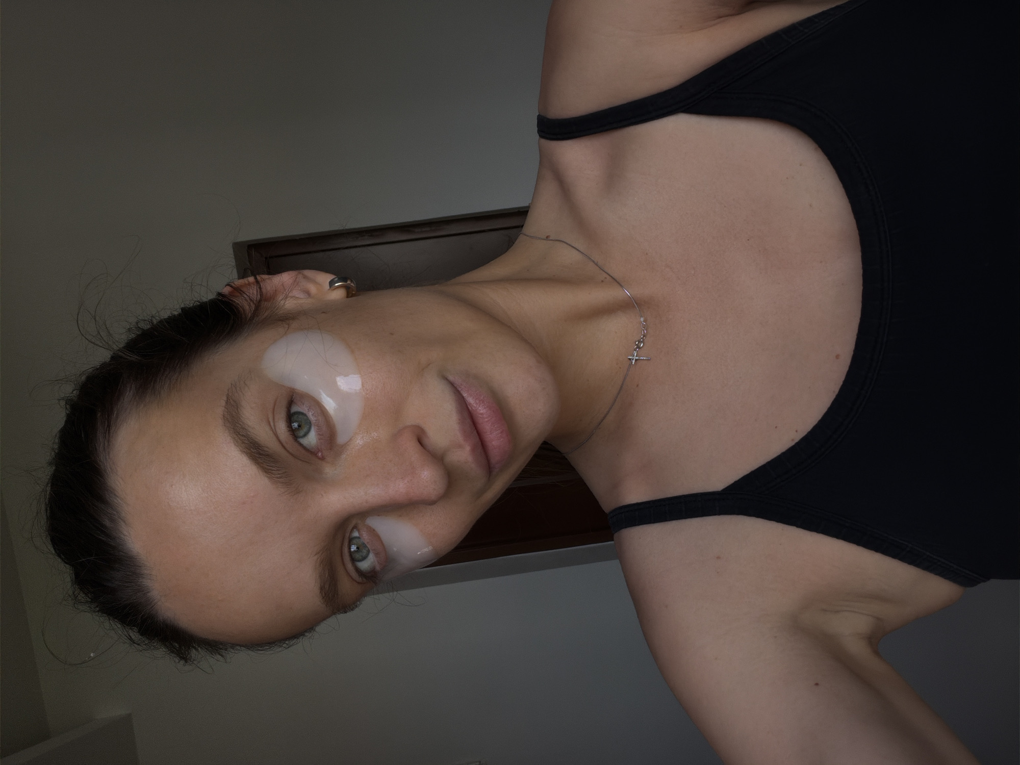 A woman with light skin and blue eyes is shown in a close-up selfie. She has dark hair pulled back and is wearing under-eye patches. She is also wearing a black tank top and a silver necklace with a cross pendant. The background is a plain wall, suggesting an indoor setting. The image does not provide any information about the location in Budva, Montenegro.