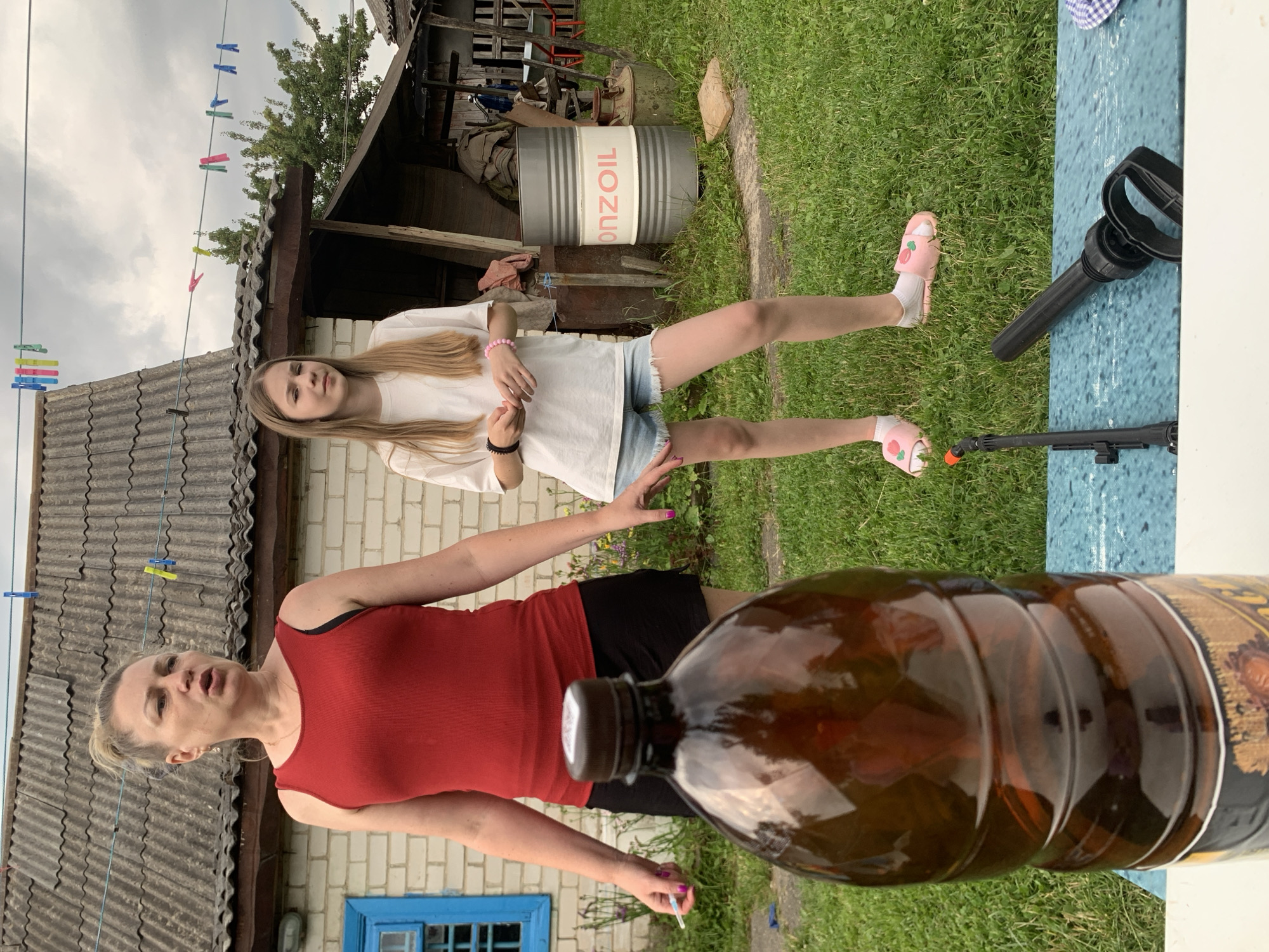In an outdoor setting, likely a rural backyard, a woman and a girl stand on a grassy lawn in front of a brick building with a corrugated roof. The woman, wearing a red tank top and black shorts, is looking to her left with her mouth open, possibly speaking. Her hair is pulled back in a bun. The girl, in a white t-shirt and denim shorts, stands to the right, looking towards the camera with a neutral expression. She has long blonde hair and is wearing pink slippers with a strawberry design. In the foreground, a large brown plastic bottle is visible, and a black object, possibly a spray nozzle or tool, lies on a blue and white patterned surface in front of it. Behind the subjects, several clotheslines with colorful clothespins are strung across the frame. A metal barrel with the label "ONZOIL" is also visible in the background, along with other miscellaneous objects and structures that suggest a utilitarian area. The overall scene appears to be a casual, possibly domestic, environment.