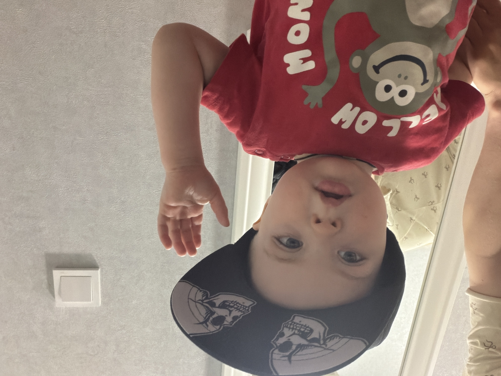 This image captures a baby viewed from an upside-down perspective, looking directly into the camera. The child has light skin, blue eyes, and slightly parted lips. They are wearing a red t-shirt or onesie featuring an upside-down grey monkey graphic and the partially visible, upside-down text "MONKEY". A black baseball cap with two skull designs on its brim covers the baby's head. A partial view of an adult's arm and hand is visible in the top right corner, suggesting the adult is holding the baby in this playful position.
The background features a light grey or beige textured wall. A white, rectangular electrical light switch is visible on the wall to the left of the baby. Behind the baby's head and to the right, a white door frame or trim is present, with a light-colored fabric featuring a subtle pattern visible beyond it, possibly part of bedding or a blanket. The scene appears to be set indoors, likely within a home in Votkinsk, Russia. The baby's expression is neutral and curious, indicative of a playful and engaging moment.