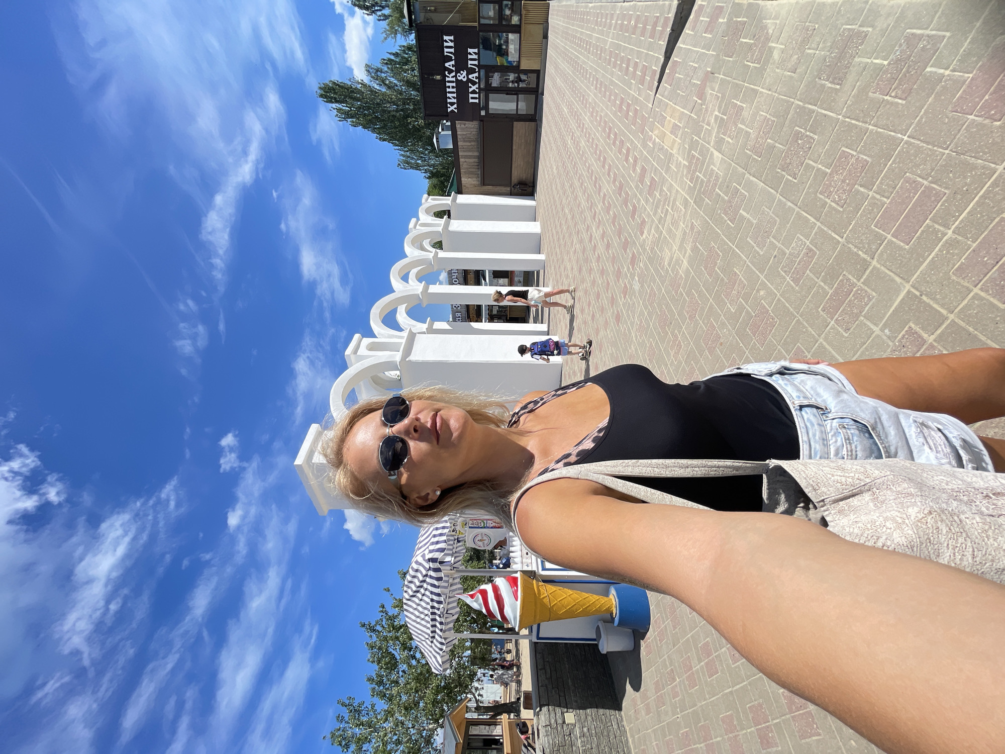 An outdoor, eye-level, wide shot captures a woman taking a selfie while standing on a paved plaza. The woman, who is facing to the right and looking slightly upward, has blonde hair and is wearing sunglasses, a black tank top, and denim shorts. A patterned tote bag is slung over her left shoulder.
Behind her, a decorative white archway with multiple arches spans the plaza. To the left, a large, three-dimensional ice cream cone statue stands beneath a striped umbrella. Further in the background, a building with text that appears to be in Russian is visible, with trees and a blue sky with wispy clouds overhead. Two children are walking in the distance, one on a scooter. The plaza is paved with interlocking rectangular bricks in a light brown and grey pattern, with shadows cast by the sun.