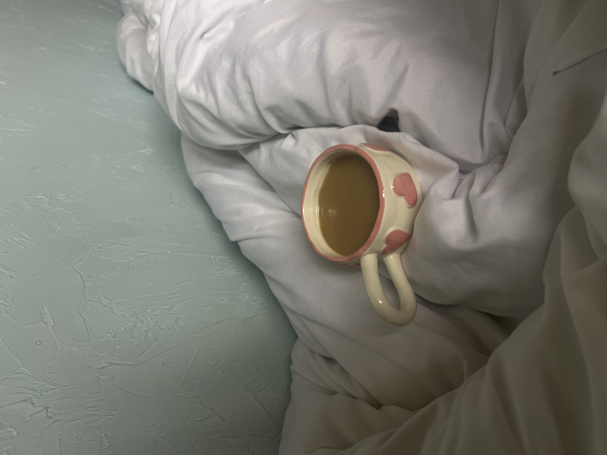 A top-down shot shows a mug of coffee sitting on a white duvet. The mug is light beige with pink heart-shaped decorations. The duvet is rumpled. To the left of the duvet is a wall painted with a light teal color. The location is in the city of Unknown, Unknown.