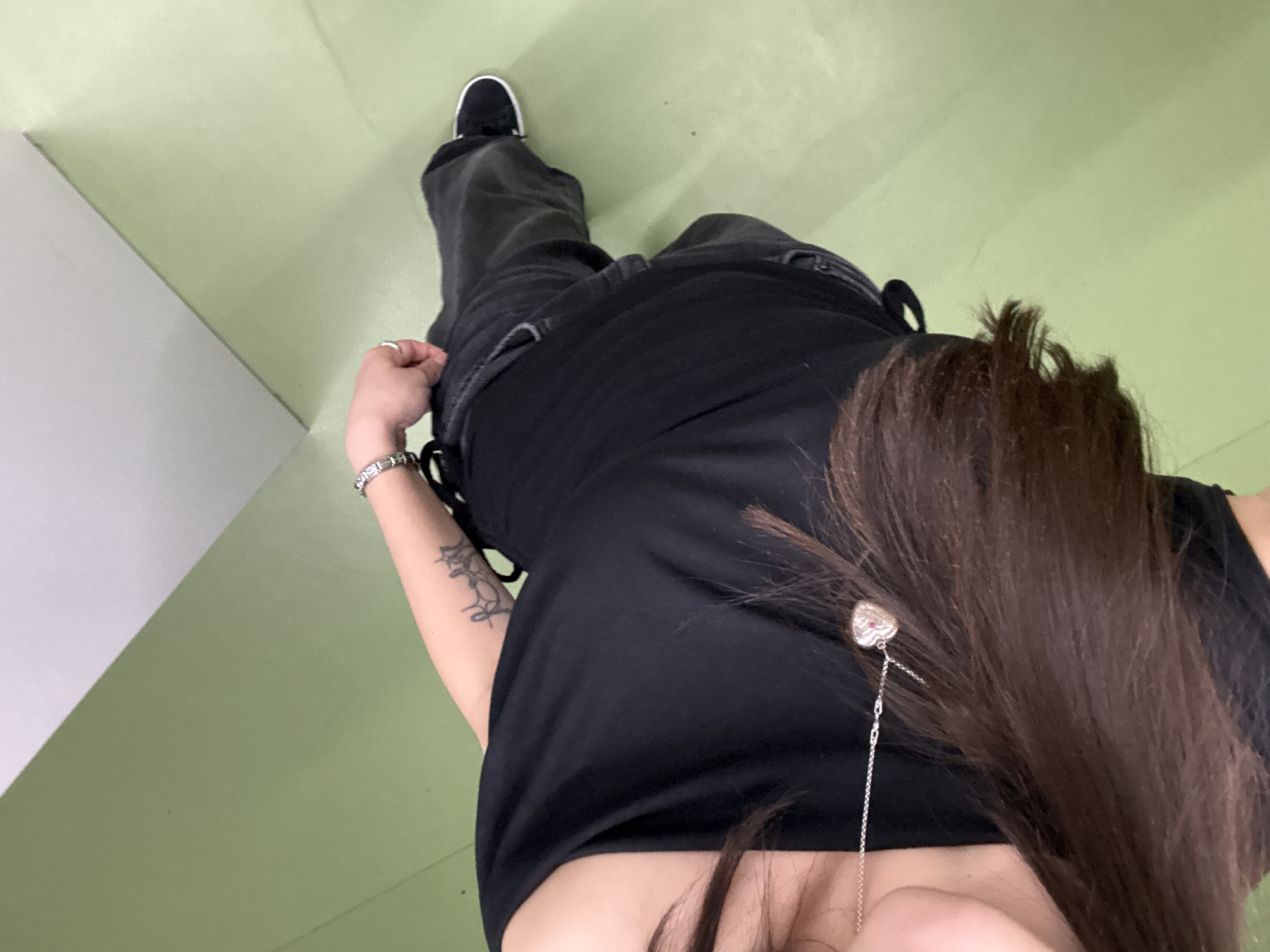 The image displays a high-angle overhead view of a person standing indoors. The individual is wearing a black sleeveless top, dark-colored trousers, and black sneakers with white soles. Long, dark brown hair partially covers the left shoulder and chest. A silver-toned necklace with a heart-shaped pendant, featuring a red stone, is visible on the chest. The person's right forearm exhibits a dark-colored, possibly calligraphic, tattoo. A silver-toned bracelet is on the right wrist, and a silver-toned ring is on the ring finger of the right hand, which is lightly grasping the upper edge of the trousers. The floor is a uniform light green, meeting a light grey or white wall at an angle in the background. This scene is located in Yerevan, Armenia.