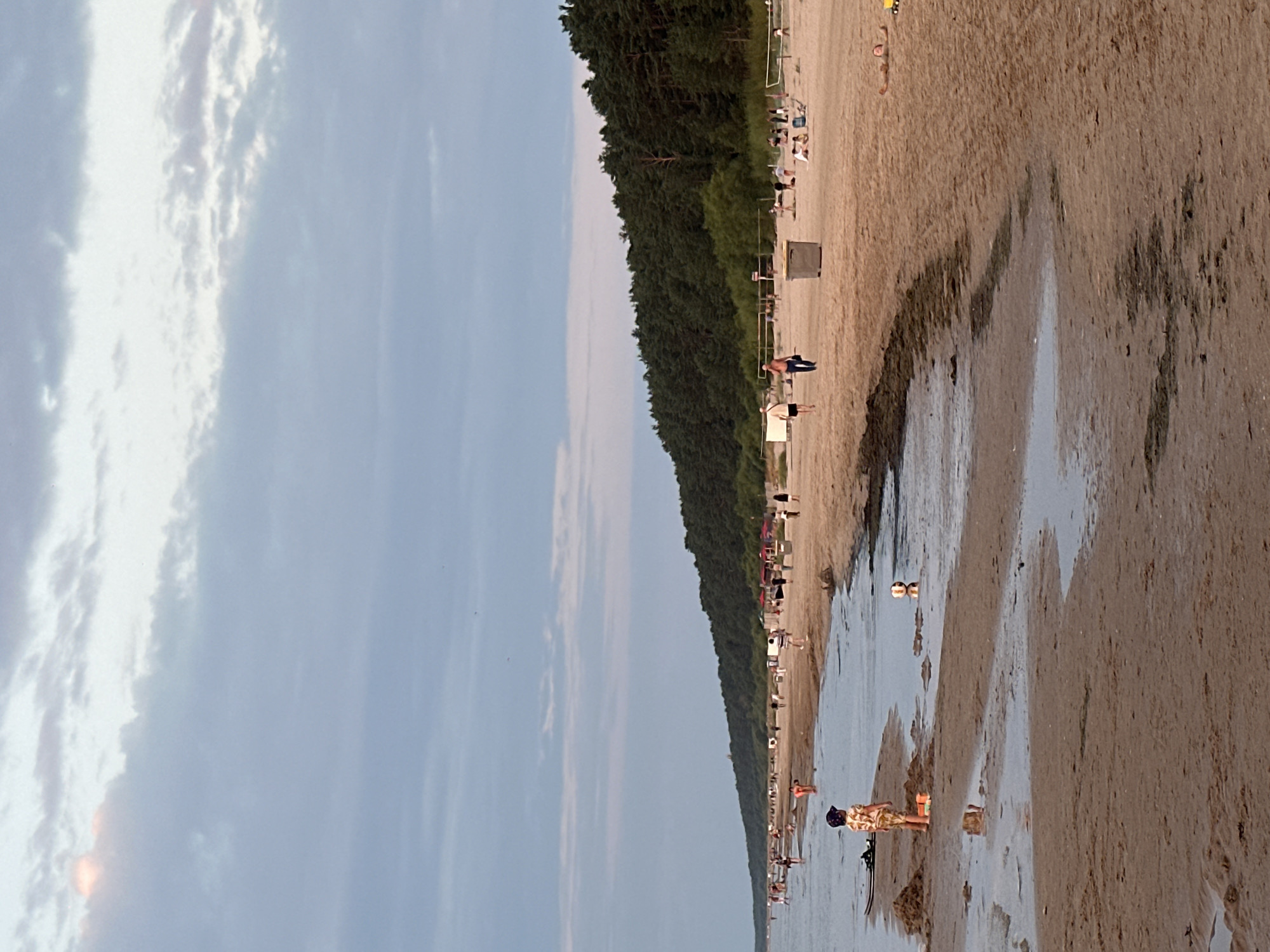An aerial, ground-level, full shot shows a wide, sandy beach in Jurmala, Latvia, with a forest bordering the sand dunes and the sea on the left. The sky is overcast with pale blue and gray clouds, with a hint of pink from the sunset in the distance. Several people are spread out across the beach, some walking along the shoreline, others relaxing on the sand or playing near the water's edge. A young child in a patterned shirt and shorts stands near the water, with sand toys around them. A soccer ball floats in the shallow water. Further down the beach, a volleyball net is set up, with a few people playing. Beach chairs and umbrellas are scattered across the sand, and a trash bin is visible near the volleyball court. The sea is calm, with gentle waves lapping at the shore. The overall atmosphere is one of a peaceful summer evening at the beach.