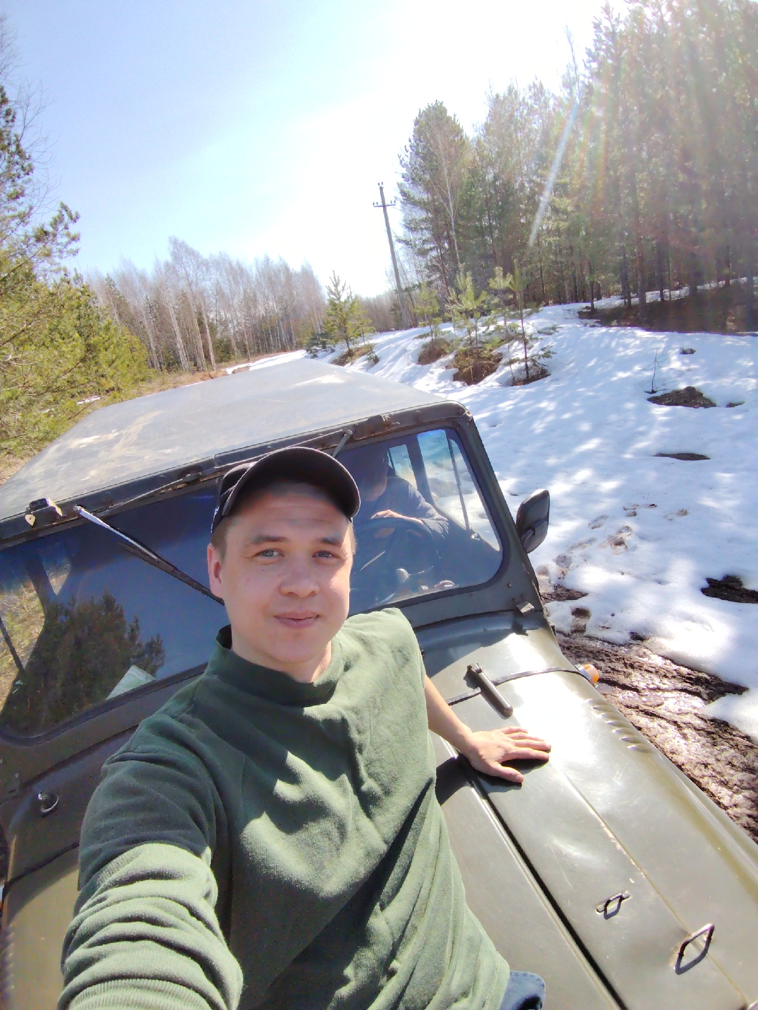 A man wearing a green long-sleeved shirt and a black baseball cap is taking a selfie, smiling slightly at the camera. He is positioned on the front of an olive-green, rugged off-road vehicle, possibly a jeep, with his left hand resting on its hood. Another person is partially visible in the driver's seat of the vehicle.
The scene depicts an outdoor, natural environment, characteristic of a forest or wooded area in Марковский, Russia. The ground is partially covered in melting snow, mixed with visible patches of mud and dirt, suggesting a transition season like late winter or early spring. The landscape features both bare deciduous trees and numerous evergreen trees, forming a dense forest in the background. A dirt track or unpaved road, suitable for off-roading, winds through the snowy and muddy terrain. A wooden utility pole with power lines is visible on the left side, slightly set back into the forest.
The image is captured in bright daylight under a clear sky, indicated by the strong sunlight, visible lens flare, and distinct shadows. The overall atmosphere suggests an adventurous outing or leisure activity in a scenic, rural, and still somewhat snow-covered landscape. There is no visible text in the image.