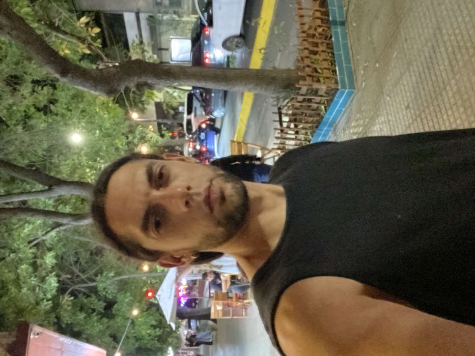 A male individual in his late 20s to early 30s, with dark hair pulled back and a short beard, wearing a black sleeveless shirt and a hoop earring in his right ear, is positioned in the foreground taking a selfie. He gazes directly at the camera.
The scene depicts a nighttime urban street in the Palermo neighborhood of Buenos Aires, Argentina, characterized by active outdoor dining and parked vehicles. To the immediate right of the individual, a tiled sidewalk transitions to a yellow-painted curb bordering a street. A tree trunk rises from a planter encircled by a low wooden slatted fence and light blue/turquoise tiles.
In the mid-ground, a vibrant outdoor café area is visible to the left, featuring multiple wooden tables and chairs, illuminated by overhead string lights. A large white umbrella with a "Heineken" logo partially obscures patrons. Several blurred figures are discernible, some seated at tables. Further down the street, a red light or illuminated sign is visible.
The background shows a street with multiple parked cars, including a blue sedan, a dark grey sedan, and a white sedan. Tall trees with dense foliage are interspersed along the street, which is illuminated by streetlights. The environment suggests a commercial or mixed-use area bustling with evening activity.