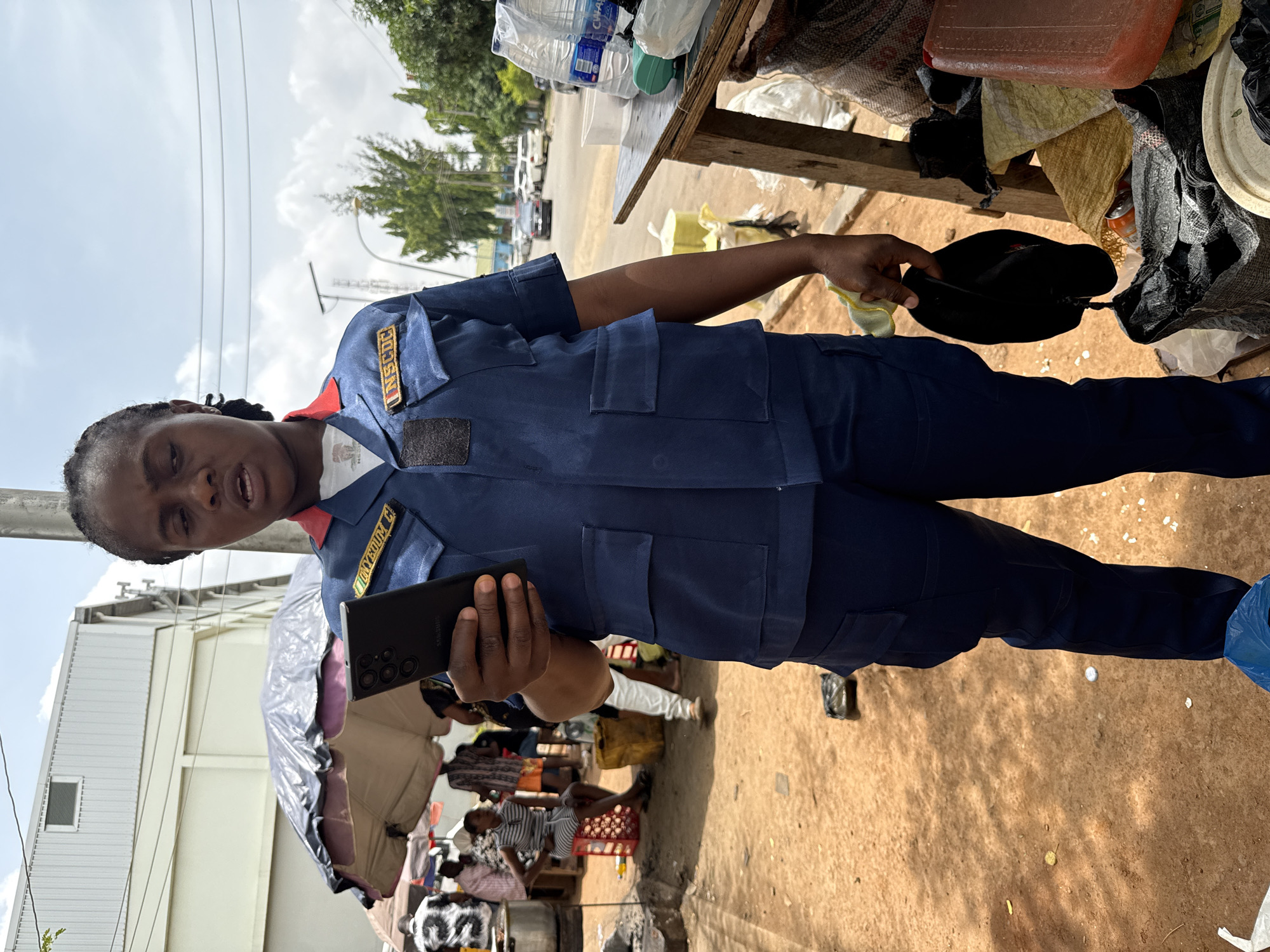 A woman in a blue uniform with "NSCDC" and the name "OYEDUNM" on the patches is holding a black mobile phone in one hand and a black cap in the other. She appears to be speaking. Behind her is a cluttered area with a table, various containers, and what appears to be a market scene with people sitting or resting. The location appears to be outdoors, on a dirt or sandy surface. The image was taken during daylight hours. The city is Abuja, Nigeria.