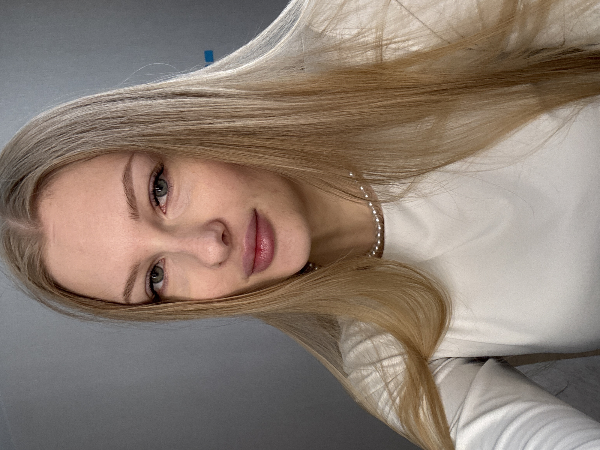 The image presents a close-up portrait of a young woman with long, straight, ash-blonde hair flowing over her shoulders. Her head is tilted to her left, with her light green or hazel eyes looking directly towards the camera. She has soft pink lips and fair, clear skin. She is wearing a simple, high-necked white top and a delicate pearl necklace around her neck. The background is a plain, light grey wall, suggesting an indoor setting, possibly in Perm, Russia. A faint blue rectangular mark is visible on the wall in the upper left area of the original frame. The lighting is soft and even, highlighting her features. The woman maintains a calm and serene expression. No specific activity is depicted other than posing for the portrait. There is no visible text in the image.