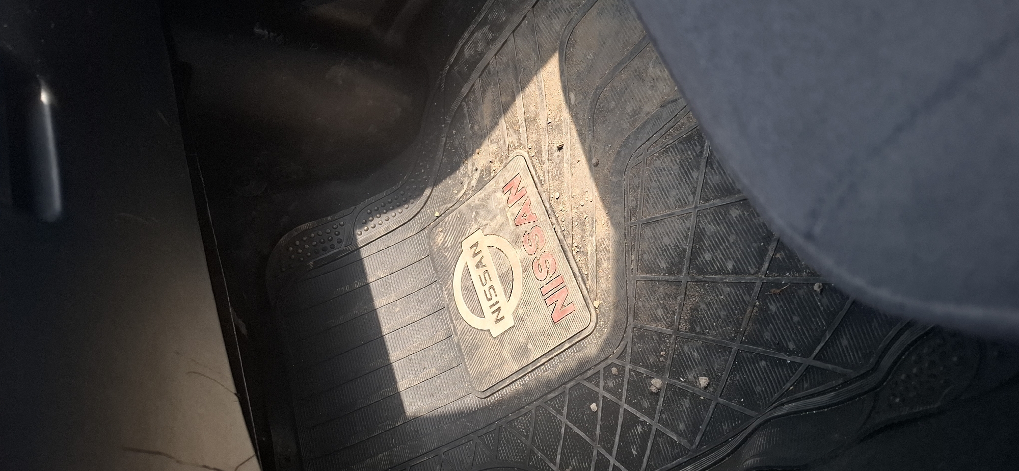 The image shows a close-up, high-angle view of a dusty black Nissan all-weather floor mat in what appears to be the driver's side footwell of a vehicle. The mat has a textured surface with raised ridges and a diamond-shaped pattern in some areas. Centered on the mat is a rectangular inset with the "NISSAN" logo embossed in a circular design and also spelled out in red lettering below it. The mat is covered in a fine layer of dust and small debris, consistent with regular use. Sunlight casts a diagonal shadow across the mat from the upper left to the lower right. The background is dark, suggesting the interior of a car. There is no visible indication of the specific city of Sychlijar, Azerbaijan, or any people.