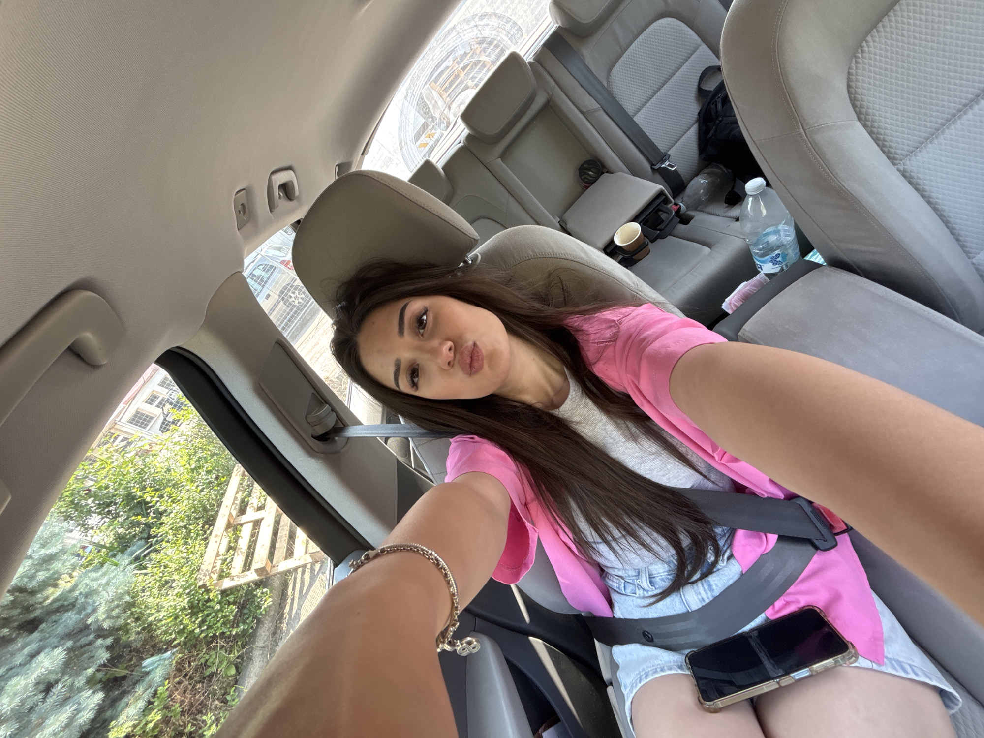 The image is a selfie taken by a young woman seated in the front passenger seat of a car, likely in Plovdiv, Bulgaria, during daytime. She has long, dark brown hair and is wearing a bright pink long-sleeved top over a light gray t-shirt and light blue denim shorts. Her left arm is extended towards the camera, revealing a silver bracelet on her wrist, while her right arm is bent at the elbow. She is making a playful "pouty" face directly at the camera. A black smartphone with a gold-toned transparent case rests on her lap.
The car's interior features light gray fabric seats and headliner. A dark gray seatbelt is buckled across her, passing over her right shoulder and lap. In the immediate vicinity, a brown paper cup is in the center console. Behind the front passenger seat, a large clear plastic water bottle with a white cap is on the floor, next to a black backpack.
Through the car windows, lush green foliage is visible. On the left side, there are dense green trees and bushes, with what appears to be part of a building with windows in the background. On the right side, through the rear window, a significant concrete structure is seen, possibly indicative of urban construction or infrastructure. The overall lighting suggests a bright, sunny day. The scene depicts a casual moment inside a vehicle.