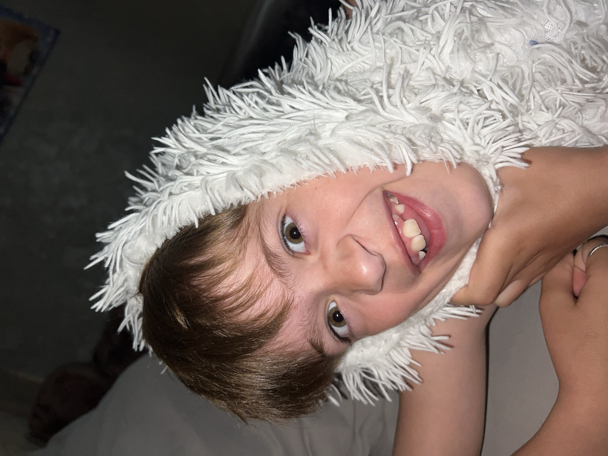 A child with light brown hair is looking at the camera with a wide smile. The child is wearing a white, fluffy head covering. An adult's hand is visible near the child's neck. The environment appears to be indoors, with a bed in the background. No information indicates the location is in Noviy, Russia.