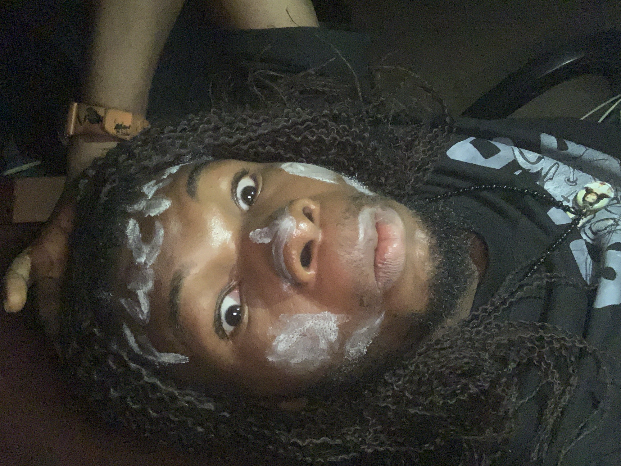 A close-up, low-angle shot of a person with dark skin and textured hair, possibly dreadlocks, lying down. Patches of white cream or paste are applied to their forehead, nose, and cheek. The person has a beard and is wearing a black T-shirt with a light-colored design. A beaded necklace with a pendant is visible. A wristband is on one wrist. The environment is dark, and the location is not discernible as Onitsha, Nigeria without further context.