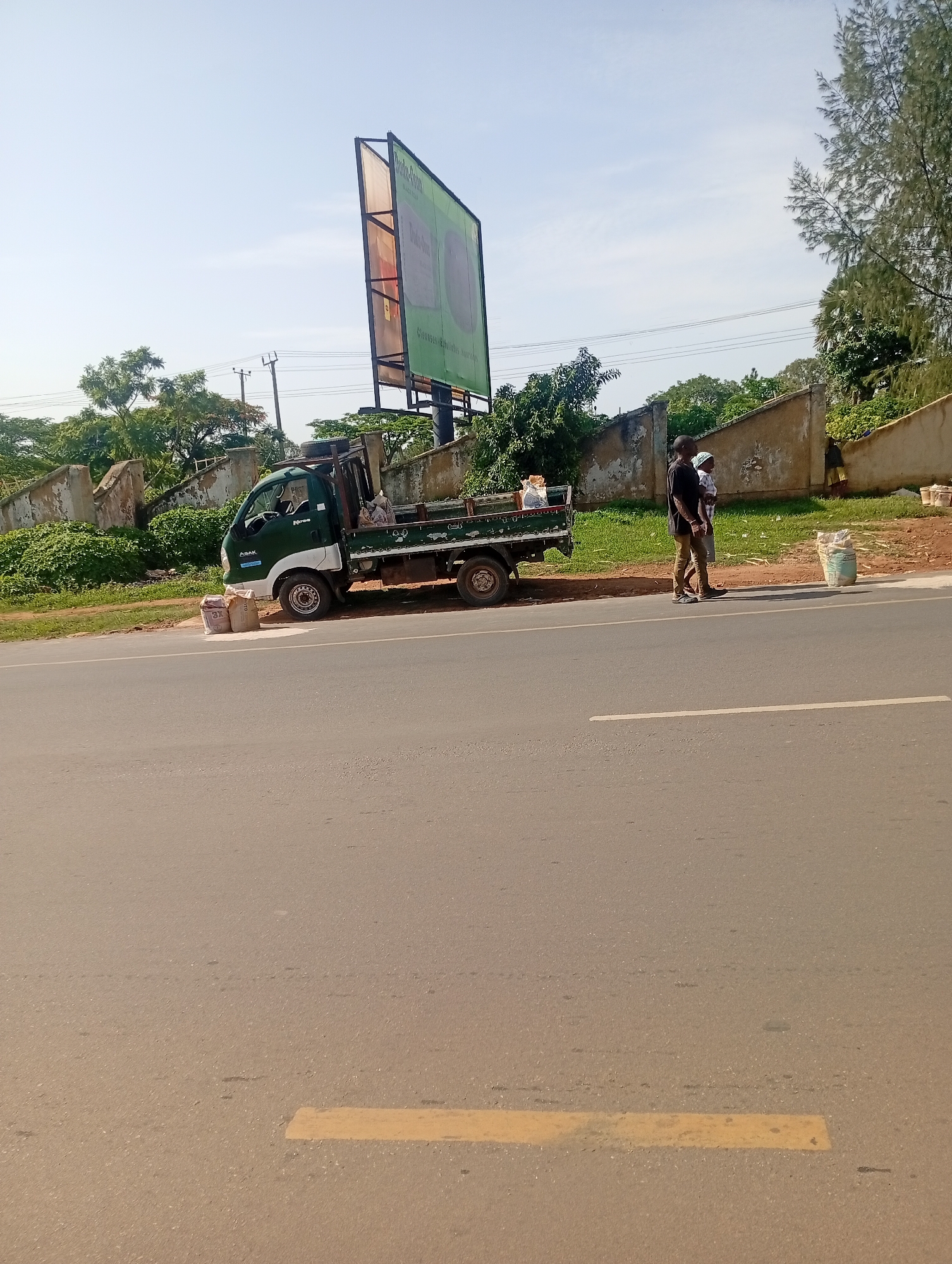The image depicts a roadside scene in Rafin Par, Nigeria. A dark green and white pickup truck is parked alongside a paved road. Bags of what appear to be cement or building materials are stacked near the front of the truck and also further down the road. Two individuals, a man and a woman, are standing on the side of the road, facing away from the camera. The man is wearing a black t-shirt and tan pants, while the woman is wearing a headscarf and a light-colored garment. A large, green billboard stands in the background, along with some trees and a weathered wall. The sky is clear and blue.