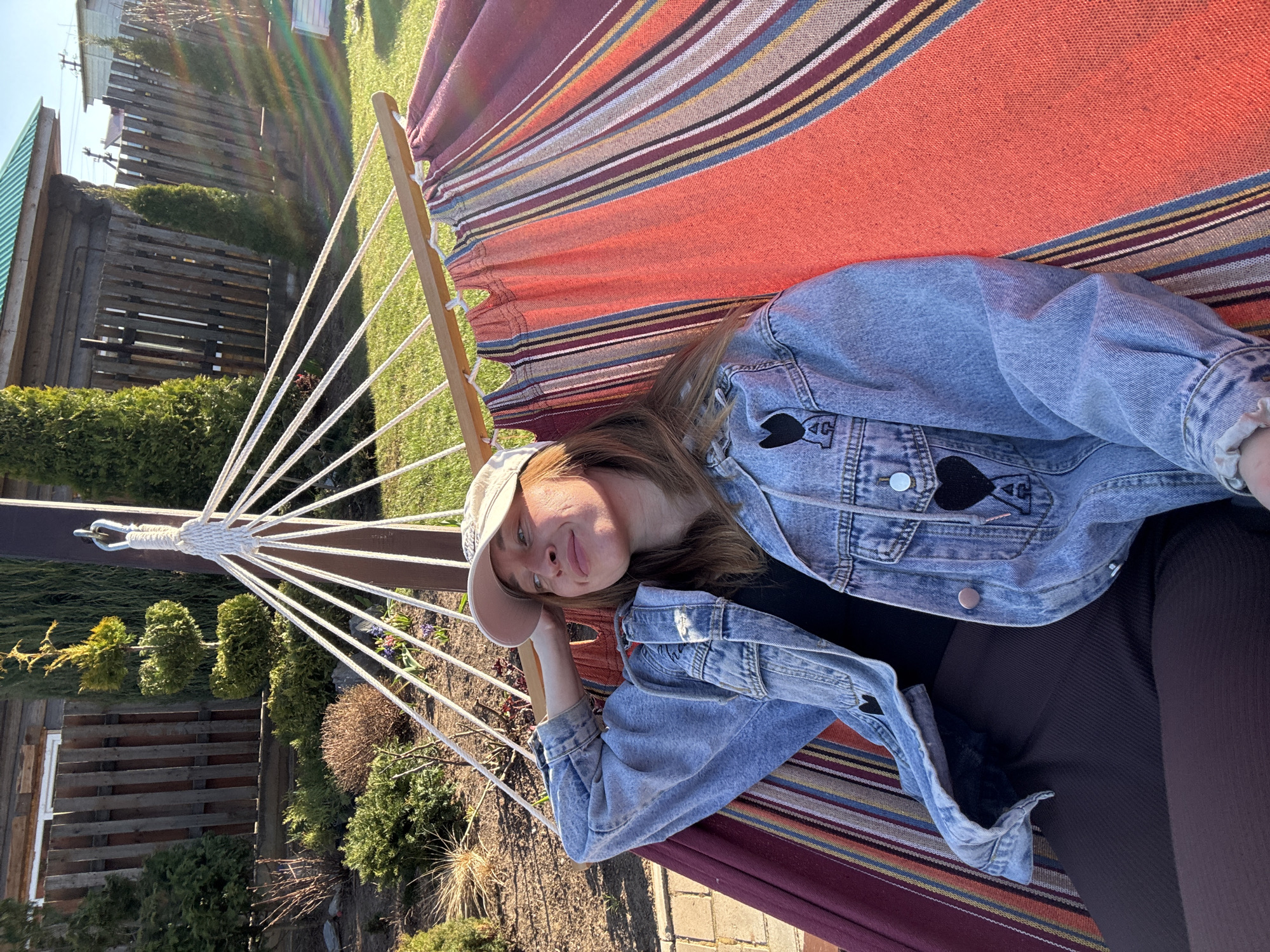 A young woman wearing a denim jacket and a baseball cap reclines in a striped hammock. The hammock is suspended outdoors, with a wooden fence and greenery visible in the background. The location is suggested to be Прикамский, Russia. The woman rests her head on her hand and gazes toward the camera.