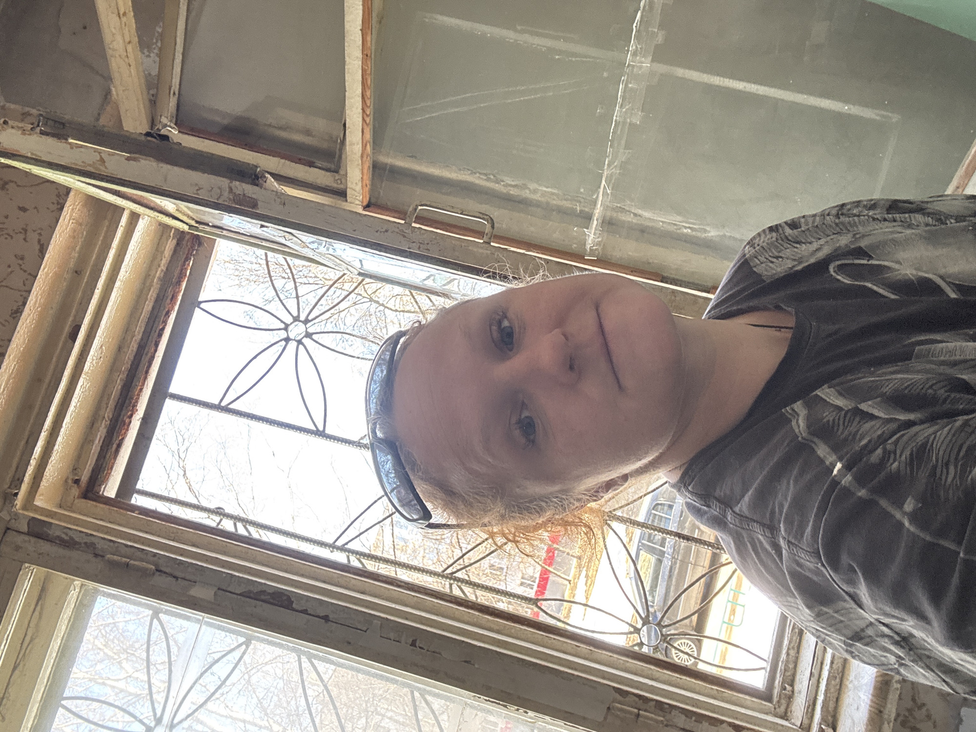 A woman with red hair and wearing sunglasses on her head is positioned in front of a window with decorative wrought iron. The window frame appears old and weathered. Through the window, a building with yellow and red elements can be seen. It is not possible to confirm the exact location as Chaikovsky, Russia from the image alone.