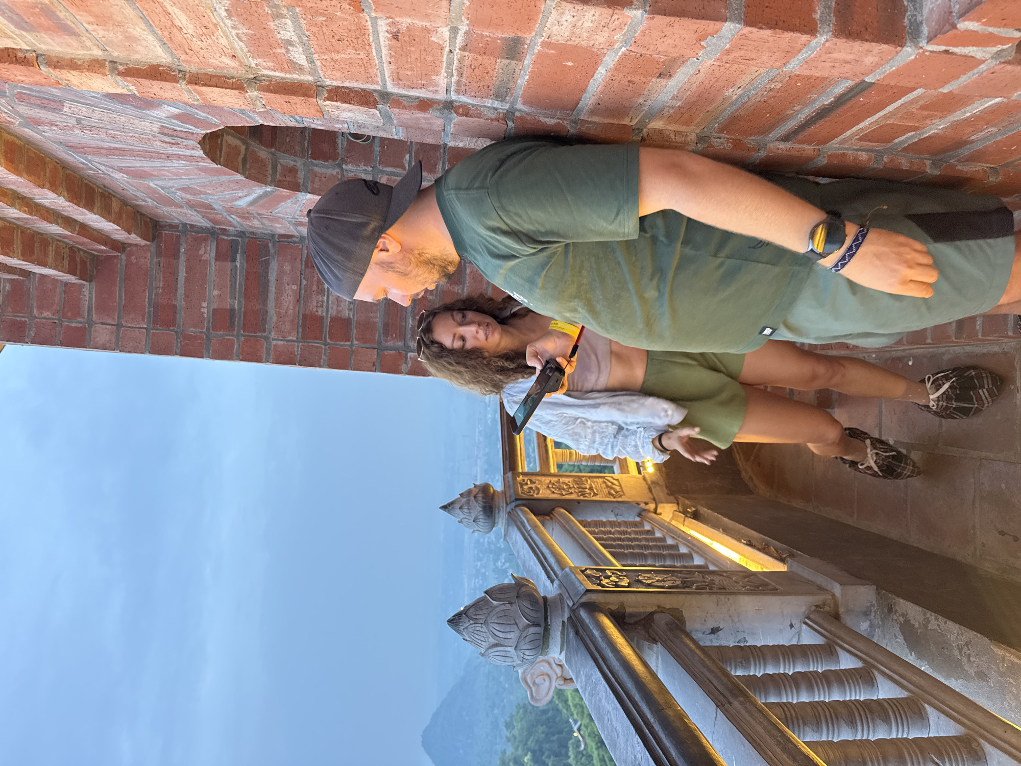 A man and a woman are standing on a brick balcony overlooking a landscape at dusk. The man is wearing a green t-shirt, green shorts, and a black baseball cap. He has a beard and is looking down at his phone, which he is holding in his right hand. A watch is visible on his left wrist. The woman is wearing a light-colored crop top and green shorts. She has sunglasses perched on her head and is also looking at a phone, which she is holding in her left hand. Her right arm is extended slightly as if gesturing or reaching out. Both individuals appear relaxed and engaged with their devices.
The setting is characterized by a brick wall with an arched opening, suggesting an old structure or architectural feature. Below the balcony, a staircase with ornate stone carvings and metal railings is visible, illuminated by warm artificial lights. The landscape in the background features rolling hills covered in lush greenery, with a hazy sky indicating either evening twilight or a slightly overcast condition. The overall atmosphere seems peaceful, with the soft light suggesting it is either early morning or late evening. The location context provided places this scene in Tây Hoa Lư Ward, Vietnam, hinting at a possibly historical or scenic tourist spot. No visible text is present in the image.