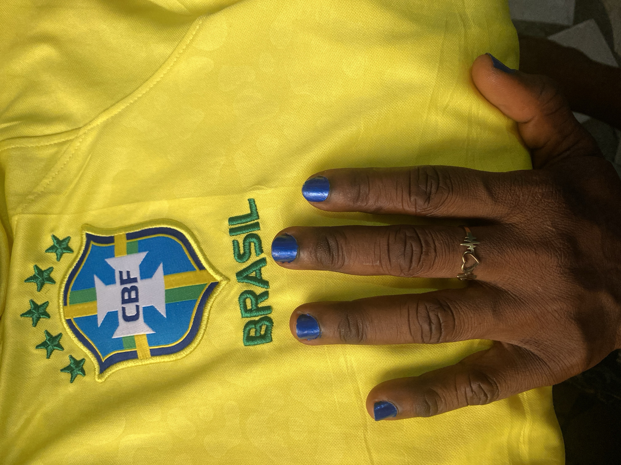 A close-up shot shows a yellow Brazil soccer jersey, emblazoned with the CBF logo and the word "BRASIL" in green. A hand with dark skin and blue painted nails rests on the shirt, adorned with a ring. The image does not provide any location context to confirm its origin or presence in Lokoja, Nigeria.