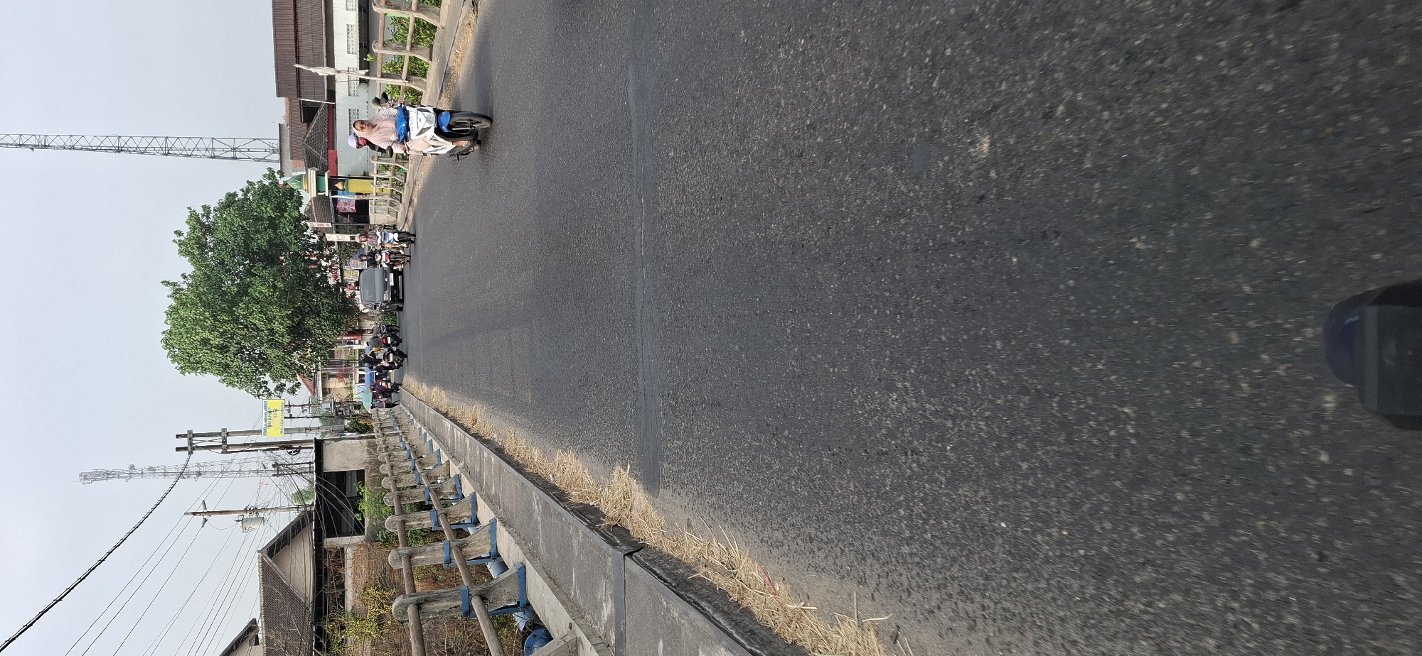 This image shows a street scene in Mranggen, Indonesia, likely on a bridge. A dark asphalt road stretches forward, with a concrete walkway and railing on the left side, lined with dry grass. Several motorcycles and a car are traveling on the road. On the right side, a white motorcycle with a blue trim is ridden by a person wearing a light pink hijab and a light-colored shirt. In the background, there are buildings, a large green tree, and communication towers. The sky is overcast.