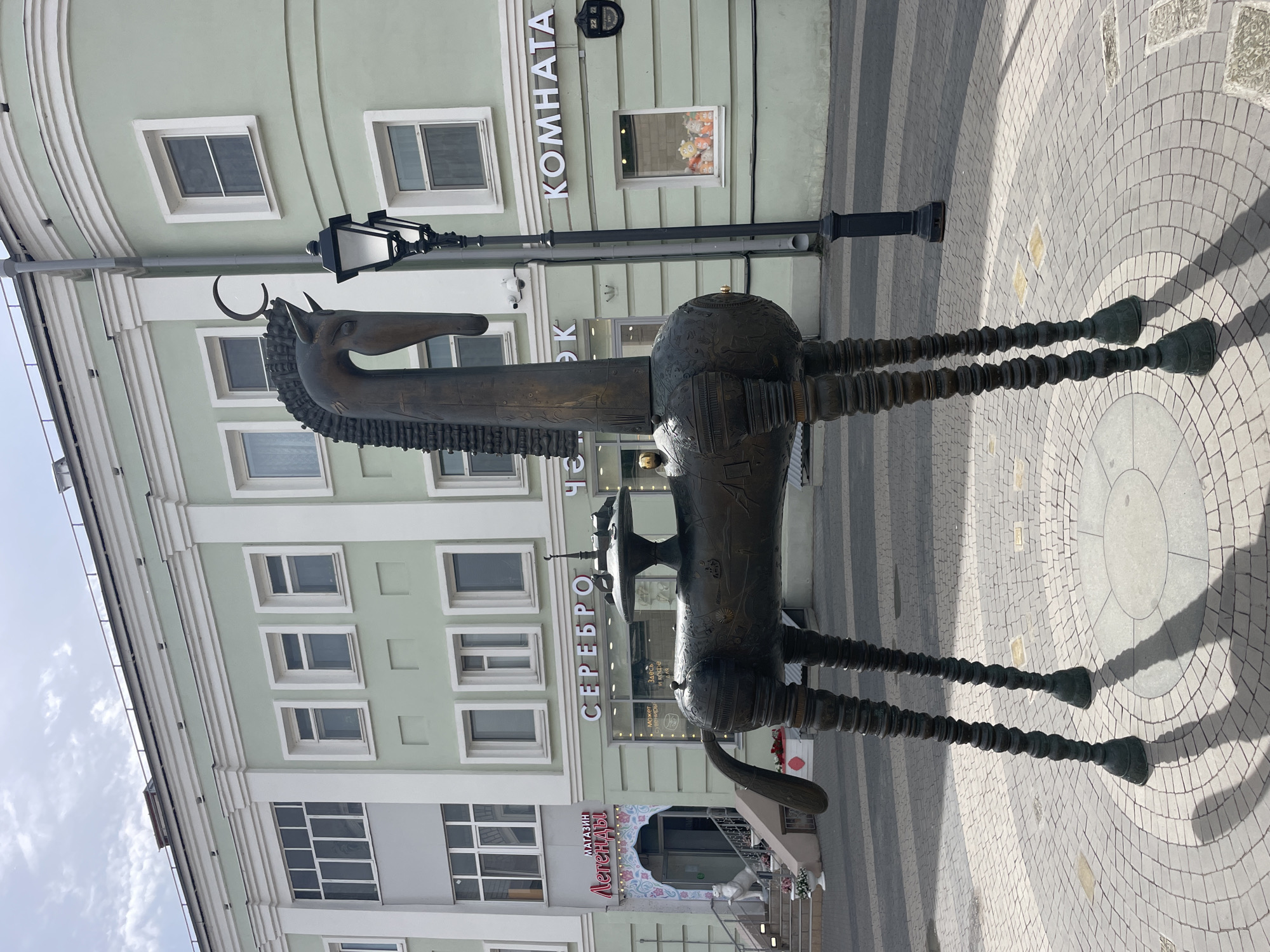 A bronze horse sculpture stands on a circular, cobblestone street in Kazan, Russia. The horse is tall and stylized, with a long neck, slender legs, and an ornate body. The statue is positioned in front of a light green building with multiple windows and signs in Russian, including "KOMNATA" and "CEREBRO." A black lamp post stands behind the horse. The street is paved with light and dark gray cobblestones. The sky is visible in the top left corner of the frame.