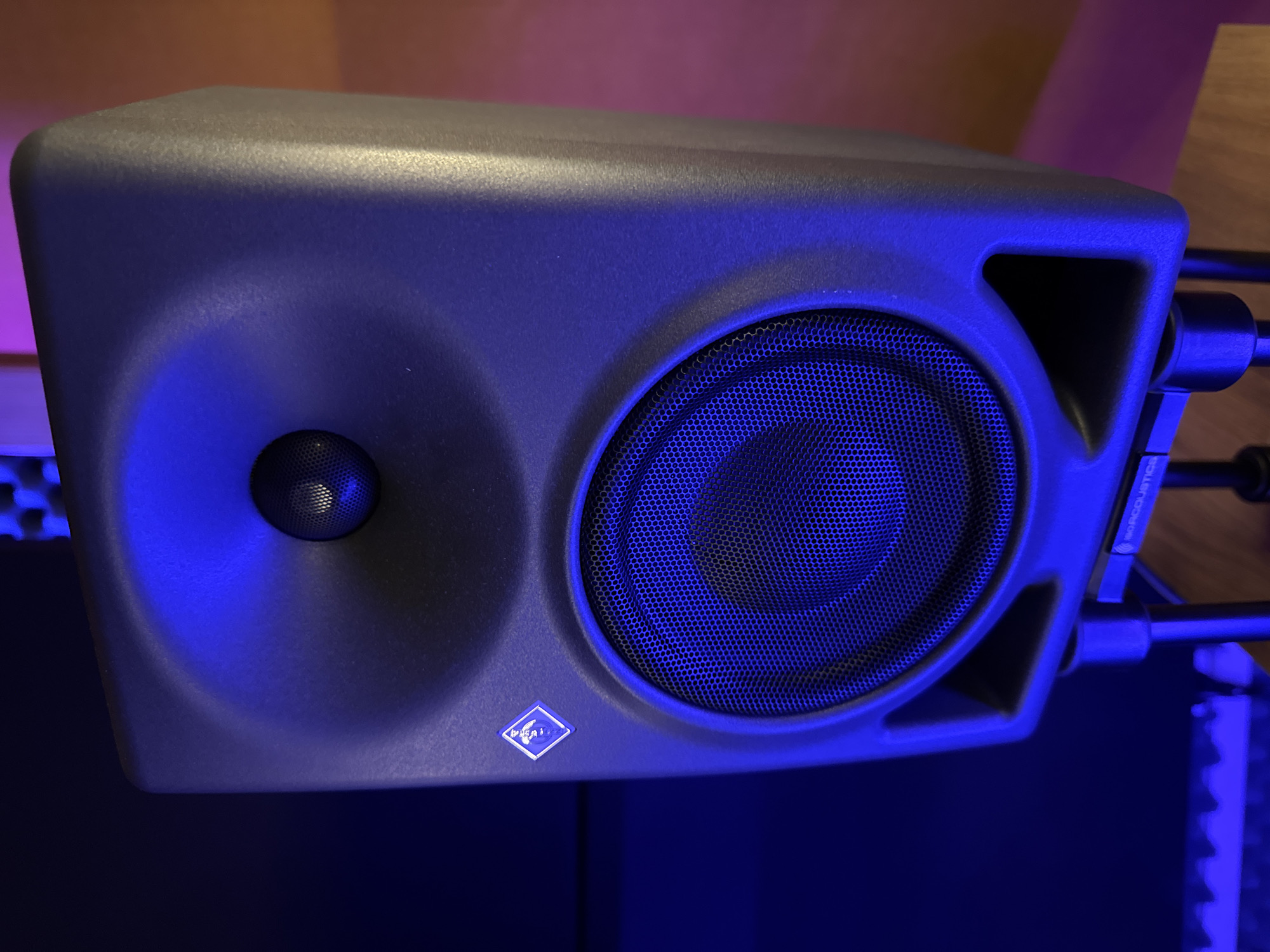 A close-up, slightly elevated shot focuses on a matte-gray Neumann KH 150 studio monitor speaker. The speaker is predominantly illuminated by blue light, with hints of purple and orange in the surrounding areas. The speaker is positioned on a desk and supported by IsoAcoustics stands. The location in the city of Plovdiv, Bulgaria cannot be determined from the image.