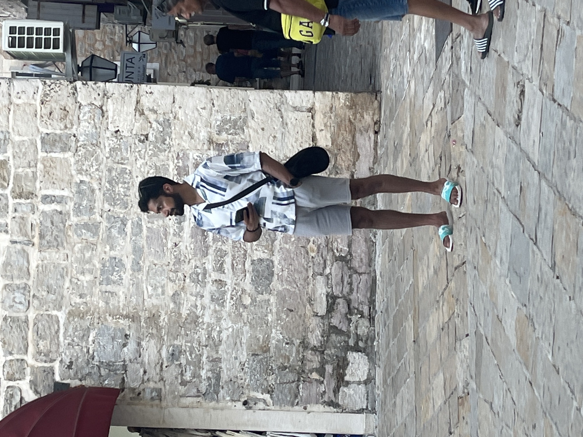 A man stands in front of a stone wall in Budva, Montenegro, looking to his left. He is wearing a white and blue patterned short-sleeved shirt, gray shorts, and light blue and white slides. He is holding a black smartphone in his right hand and has a black crossbody bag slung over his left shoulder. The street is paved with stone tiles. In the background, another man is visible from the chest down, wearing blue shorts and black and white Adidas slides. He is carrying a bright yellow bag with "GA" visible on it. Further in the background, two men in dark shirts and shorts are walking away from the camera. There are street lamps attached to the wall, and an air conditioning unit is mounted above them. A portion of a red awning is visible on the far left of the image.