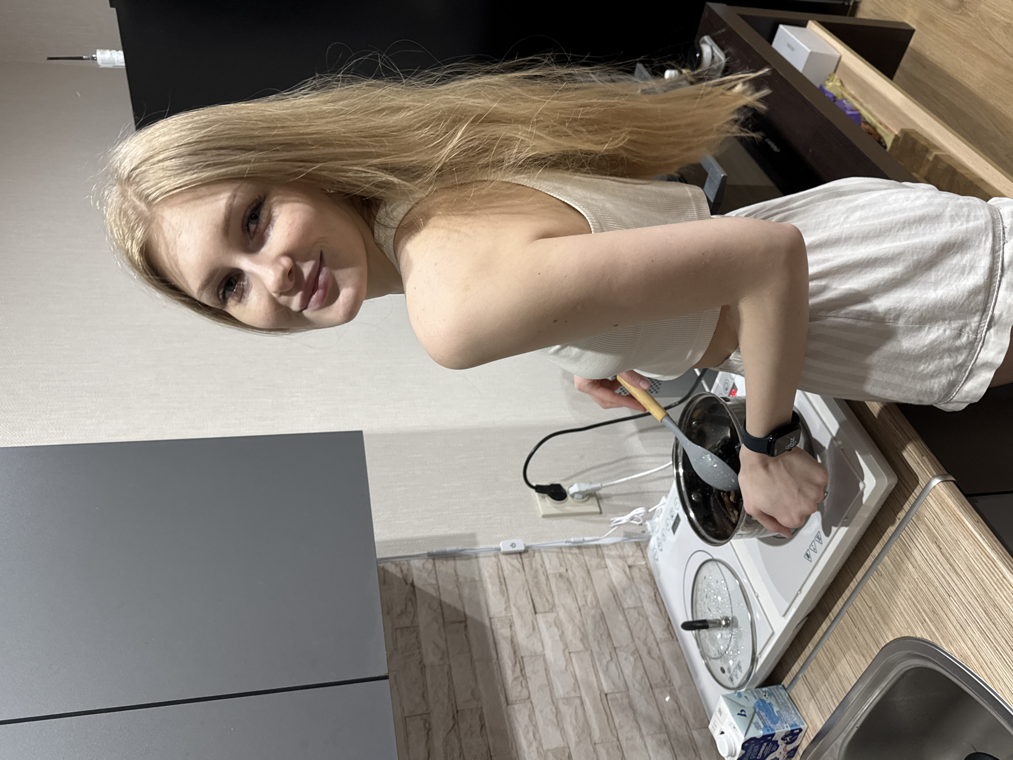 A fair-skinned woman with long, light blonde hair is standing in a kitchen. She is wearing a light-colored tank top and shorts. She is positioned at an electric stovetop, stirring the contents of a pot with a wooden-handled spoon. The stovetop is white and has a covered pot on one burner. To the side of the stove is a sink, and near the bottom of the image is a carton of milk.
The background features a gray wall, a faux brick wall, and wooden cabinetry.
Although difficult to pinpoint the exact location based on the image, the text on the milk carton may confirm that the location is in Perm, Russia.
