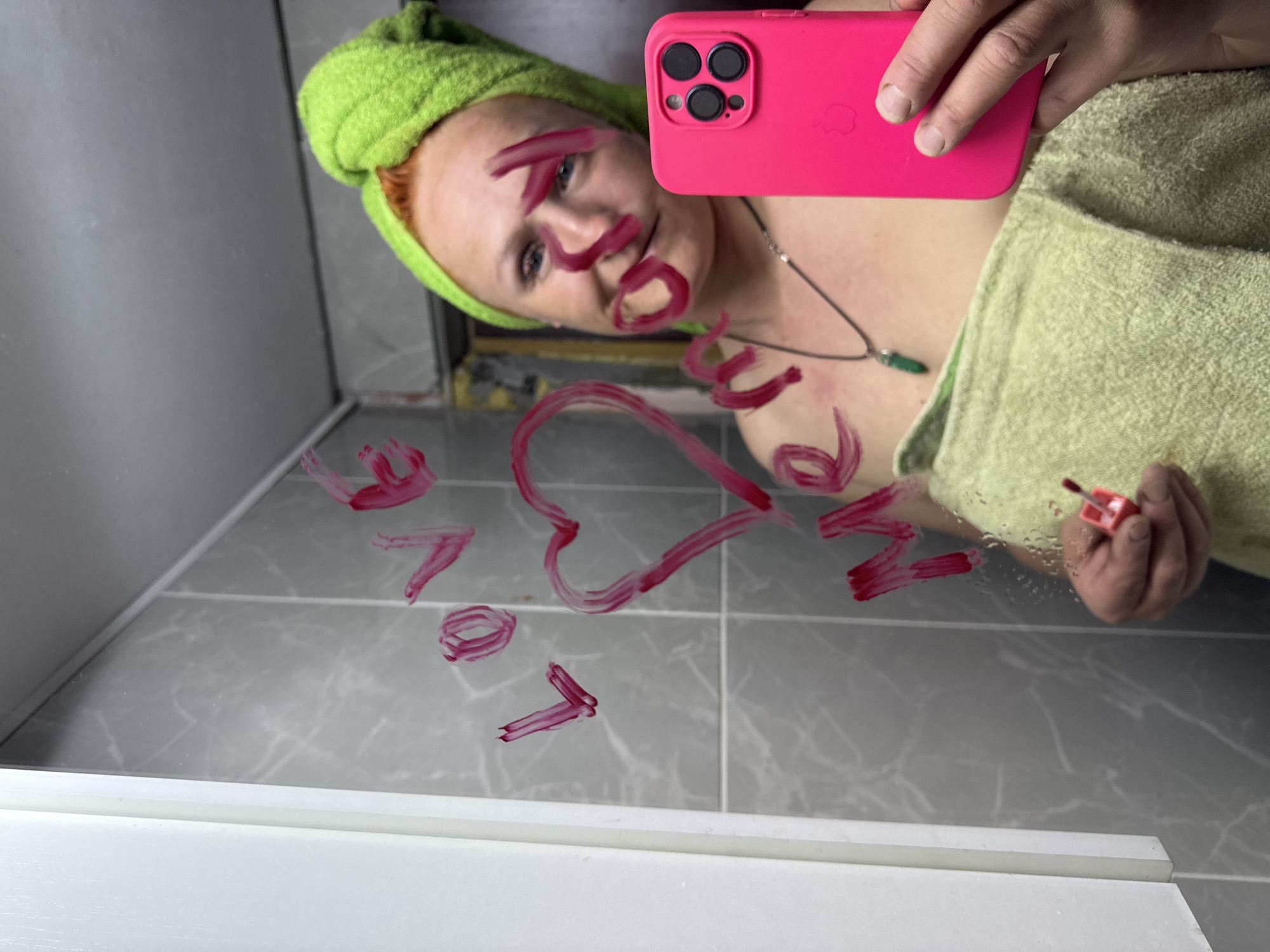 The image shows a person taking a selfie in a bathroom mirror, with the phrase "Love You" written on the mirror in what appears to be lipstick. The person has a green towel wrapped around their head and another draped over their shoulders. They are holding a pink smartphone to take the photo. A necklace with a green pendant is visible. The bathroom features gray tiles and white trim. The image was likely taken in Chaikovsky, Russia.