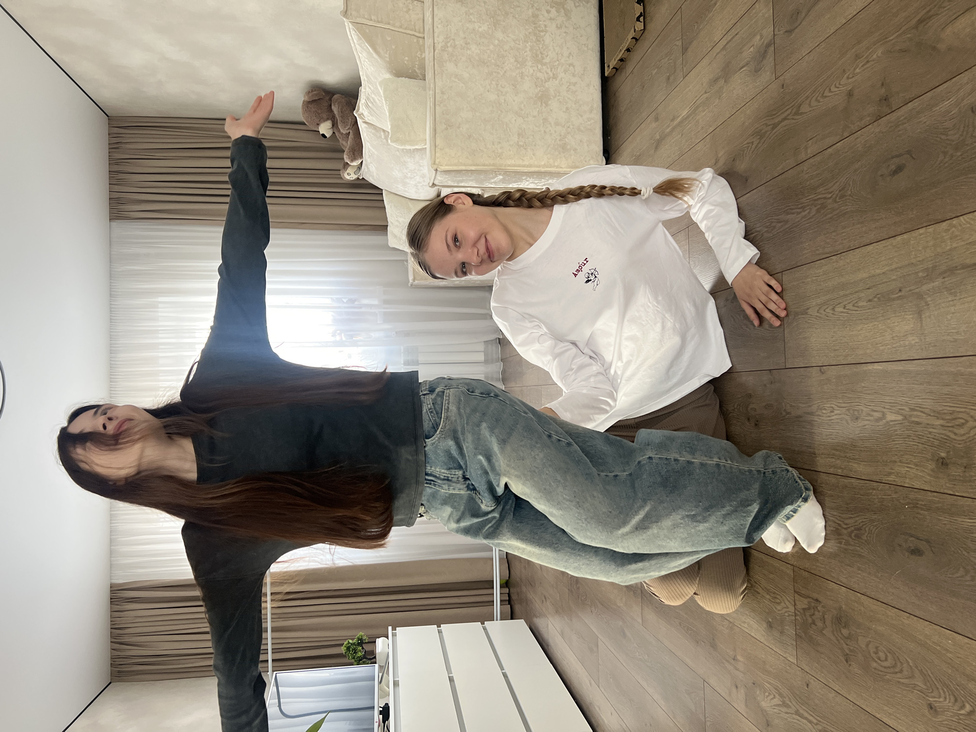 Two females are on a wood floor. One lies on her stomach, smiling at the camera, with a braid, wearing a long-sleeved white shirt. The other, wearing blue jeans and a black long-sleeved shirt, stands with one arm raised. A beige couch is near one female, and a white shelf near the other. The location is not identifiable as Votkinsk, Russia.