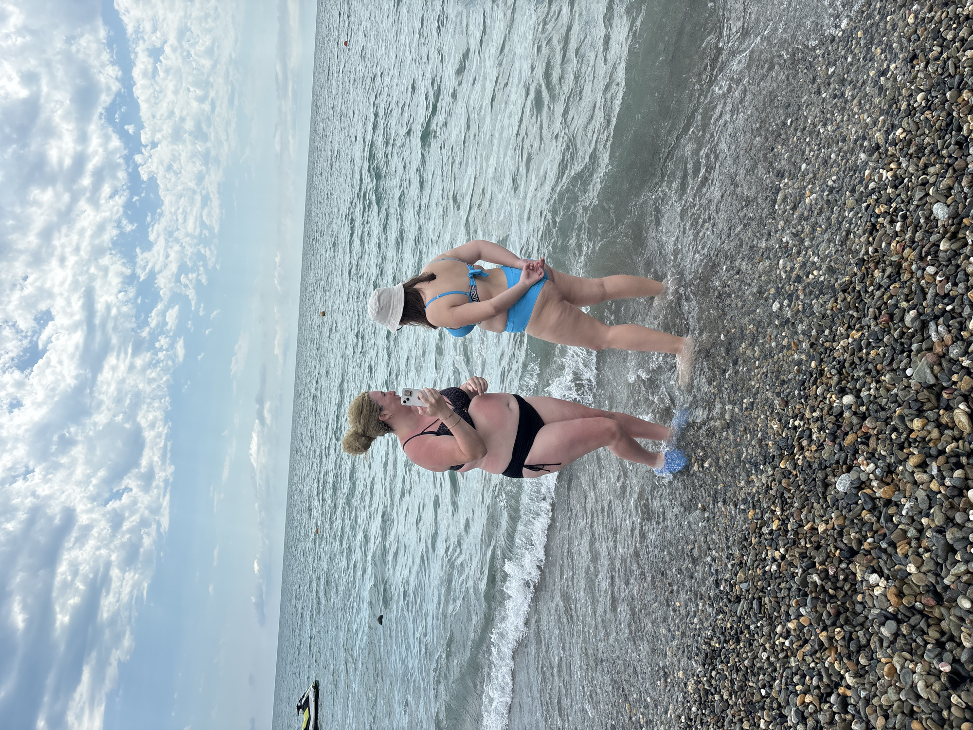 The image shows two women at a pebble beach near Sochi, Russia. The woman in the foreground is wearing a black bikini and is holding a white smartphone, appearing to take a selfie. Her hair is pulled back in a bun, and she has a slight tan. She is standing in shallow water, with her feet visible. The woman in the background is wearing a blue bikini and a white baseball cap. She is facing away from the camera, with her hands clasped behind her back, and is also standing in the water.
The scene depicts a clear day with a partly cloudy sky. The sea is calm with gentle waves lapping at the shore. The beach is covered in a variety of pebbles. The overall impression is one of a relaxed summer day at the beach. The image is oriented vertically, suggesting it was taken with a phone. There is no visible text in the image.