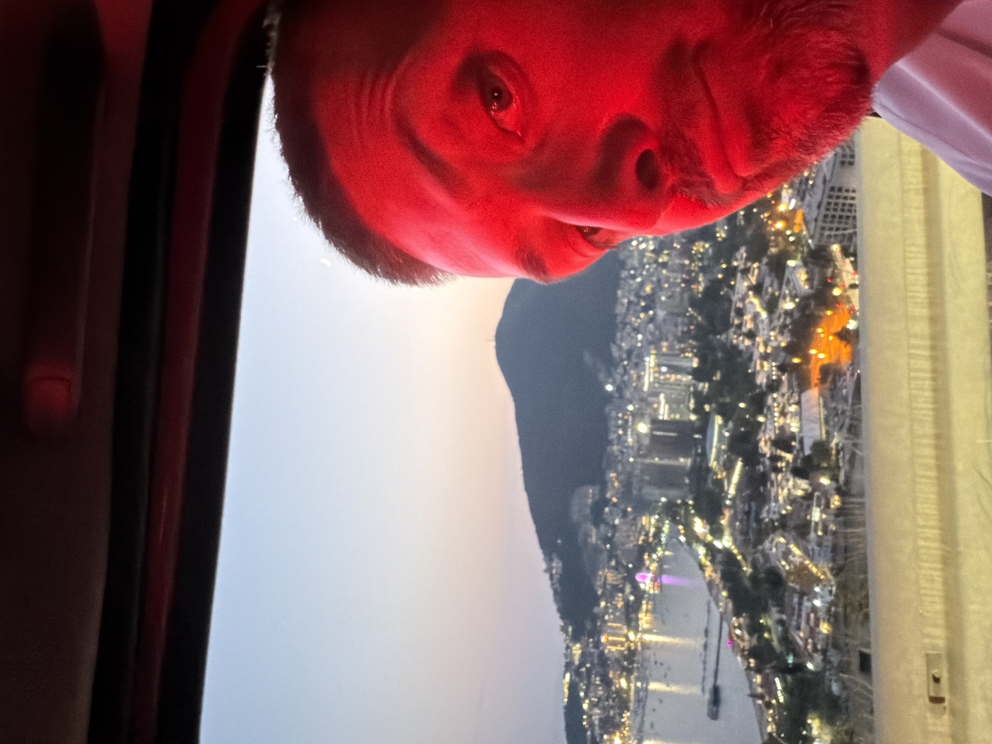 A close-up, selfie-style photograph captures a man's face, bathed in red light, from the chest up. He is looking towards the camera with his right eye visible and his mouth slightly open. The background reveals a panoramic view of a coastal city at dusk. The city lights twinkle, indicating a densely populated area with buildings and structures cascading down a hillside towards a body of water dotted with boats. A large, dark hill or mountain dominates the far background, silhouetted against a pale sky. A sliver of a crescent moon is faintly visible in the upper left quadrant of the sky. The perspective suggests the photo was taken from a high vantage point, possibly a vehicle, given the red interior visible at the top of the frame and the railing in the foreground. The specific location is identified as Košljun, Montenegro.