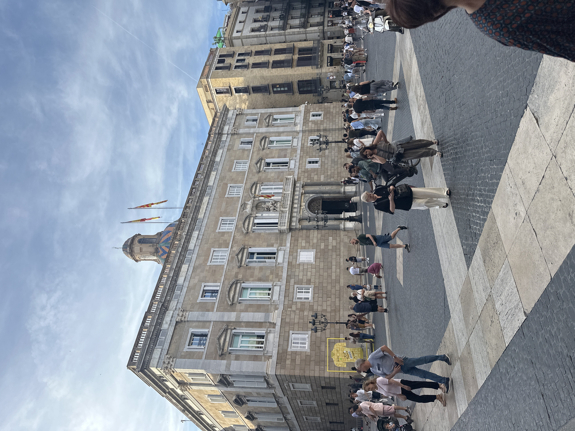 The image captures a bustling daytime scene in Plaça de Sant Jaume, Barcelona, Catalonia, Spain. In the center-left, the historic Palau de la Generalitat de Catalunya dominates the frame, a multi-story stone building with classical architectural elements, numerous windows, and white shutters. A prominent balcony above the main entrance features a coat of arms and two flags, one red and yellow striped (Catalan), the other red, yellow, and red (Spanish). On the roofline, a balustrade is visible, and behind it, a distinctive colorful tiled dome (blue, green, orange) and two additional flags. On the lower facade of this building, a yellow outline shaped like a cycling jersey is affixed, displaying text including "Tour de France" and "La Grande Boucle."
To the right of the main building, several other multi-story stone buildings with windows and balconies line the square. The foreground and midground are paved with a pattern of large square stone tiles in shades of grey and beige, interspersed with areas of dark cobblestones.
Numerous people are present throughout the plaza, engaged in various activities. Individuals and small groups are seen walking, standing, and conversing. Their attire suggests warm weather, consisting primarily of t-shirts, shorts, dresses, and light pants. Specific individuals include a person pushing a stroller on the far left, a woman in a striped shirt and dark pants, and a man in a grey t-shirt and jeans walking together in the midground. Another woman in a dark top and white pants walks on the left, while a woman in a green jumpsuit talks on a mobile phone near a man on a bicycle in the midground. Further right, a man in a green t-shirt and blue shorts walks, and a white scooter with a rider is visible. Several ornate black lampposts are scattered across the plaza. The sky above is partly cloudy, with a single faint contrail extending from the upper right. The lighting indicates late afternoon.