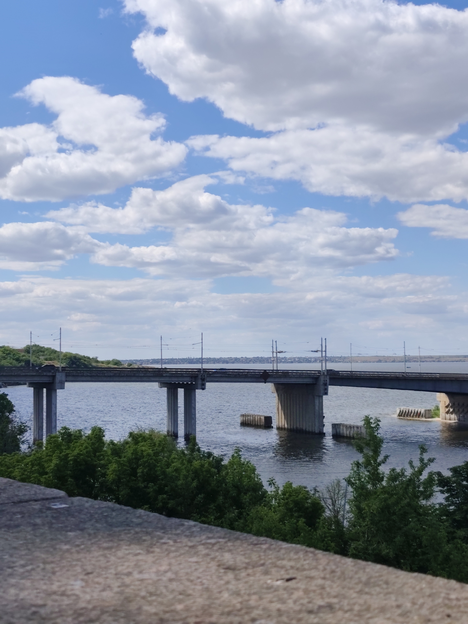 The image depicts a wide river with a large bridge spanning across it. The bridge is supported by concrete pillars and appears to be a railway bridge, indicated by the overhead power lines. The water is a deep blue, and the sky above is clear with scattered white clouds. Lush green trees line the riverbank in the foreground and midground. In the distance, a city skyline is visible on the far bank of the river. The foreground features a textured stone embankment. This scene is likely the Varvarivskyi Bridge in Mykolaiv, Ukraine, a prominent railway bridge crossing the Southern Bug river.