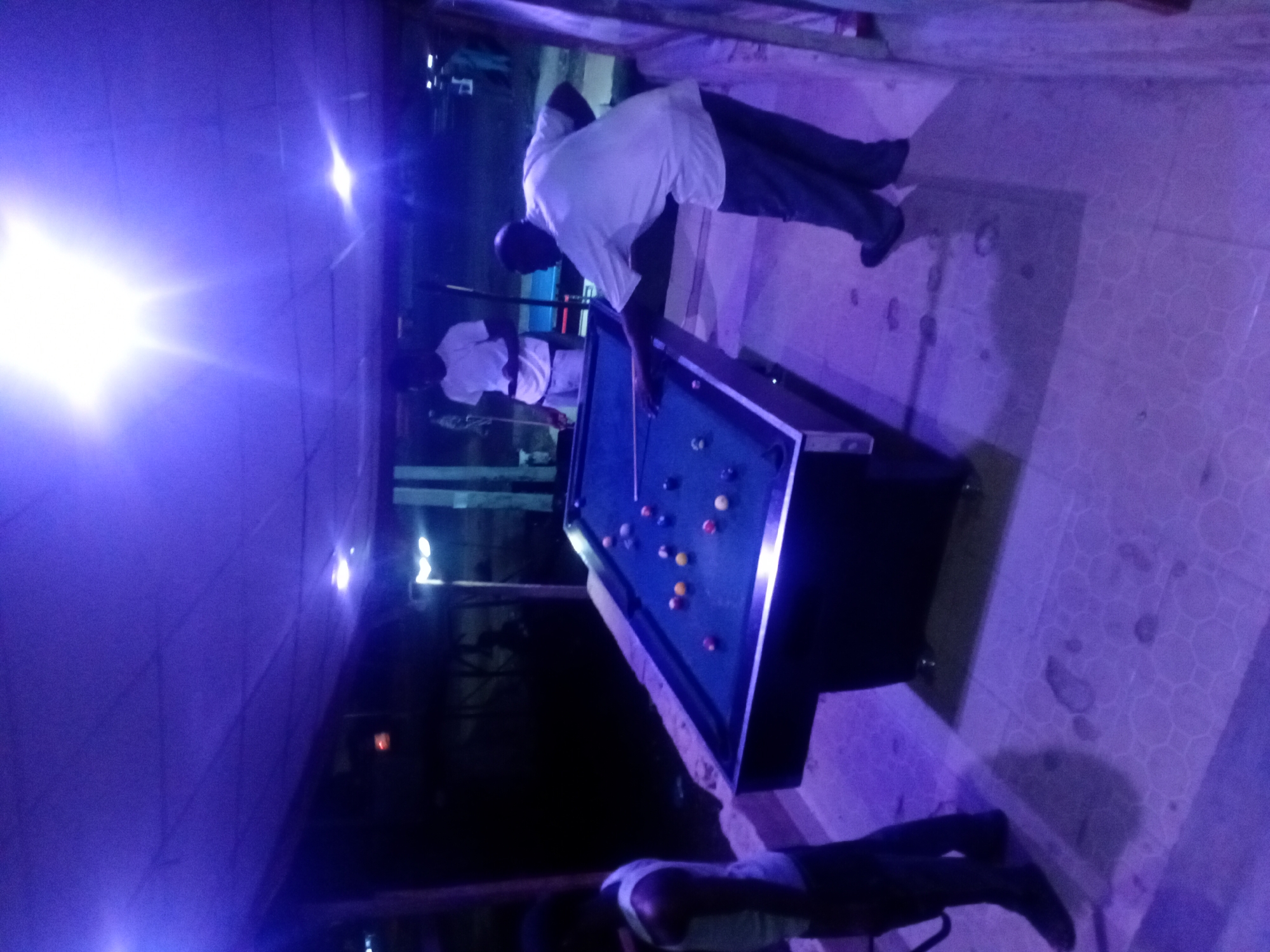 The image captures a nighttime scene of people playing pool in an indoor or covered outdoor setting, illuminated by strong artificial purple and blue light. The photo is rotated ninety degrees clockwise.
In the foreground, a man wearing a white short-sleeved shirt and dark pants is leaning over a pool table, poised to take a shot with a cue stick. His left hand is placed on the green felt surface of the table, guiding the cue. Several billiard balls of various colors (red, yellow, orange, striped, solid) are scattered on the table.
Another person, also in a white shirt, is visible in the background, near the other end of the pool table. The lower half of a third person, showing dark pants and shoes, is visible in the bottom right corner of the frame, indicating other individuals are present.
The dominant feature of the image is the intense purple and blue lighting, which casts a strong, almost monochromatic glow over the entire scene. Bright light flares, likely from overhead light sources, are prominent in the upper left and upper right areas of the original image, contributing to the ethereal and somewhat blurry appearance. The floor is tiled with a repeating circular or hexagonal pattern, visible in the areas not obscured by shadows or light. There is no visible text in the image. The atmosphere suggests a leisure activity taking place in an entertainment venue.