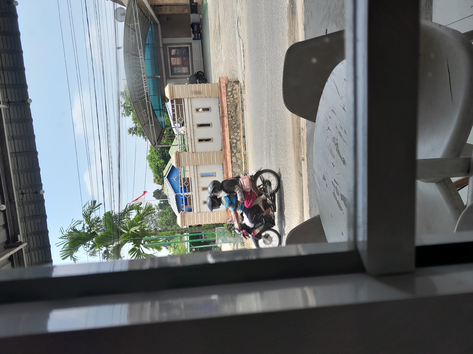 In Kotapinang, Indonesia, a man and a woman ride a motorcycle on a paved road. The man, wearing a blue and striped shirt and a black helmet, drives. The woman, in a black shirt and pink pants, sits behind him, also wearing a helmet. A pink bag is attached to the back of the motorcycle. They pass a beige, decorative wall with rectangular openings, a tall palm tree, and a building with a gray roof and a blue facade. The scene is viewed through a window with a metal frame, with a white table and chairs visible in the foreground. Overhead, a network of electrical wires crisscrosses the bright, blue sky.