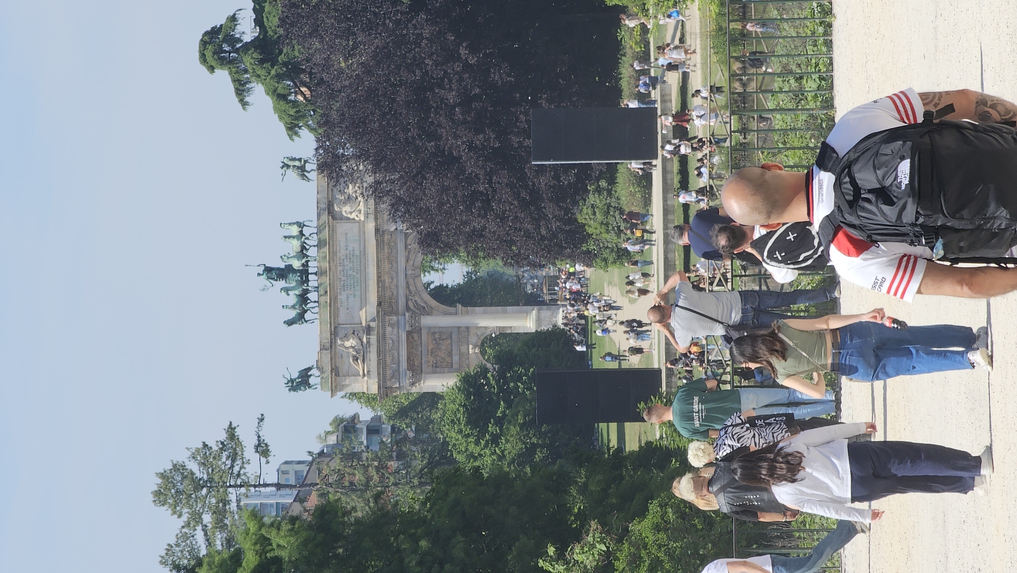 The image captures a sunny outdoor scene in Milan, Italy, featuring the iconic Arco della Pace (Arch of Peace) monument as a focal point. The arch, adorned with sculptures, stands at a distance, framed by lush greenery and trees. A crowd of people populates the area in front of the arch, walking on the concrete pavement and gathering near the grassy areas. Several individuals are visible closer to the camera, including a bald man with a backpack wearing a white shirt with red stripes and light colored shorts. There is a group of other people of various ages and clothing styles are interspersed within the scene. In the background, buildings and a clear blue sky are visible, completing the setting.