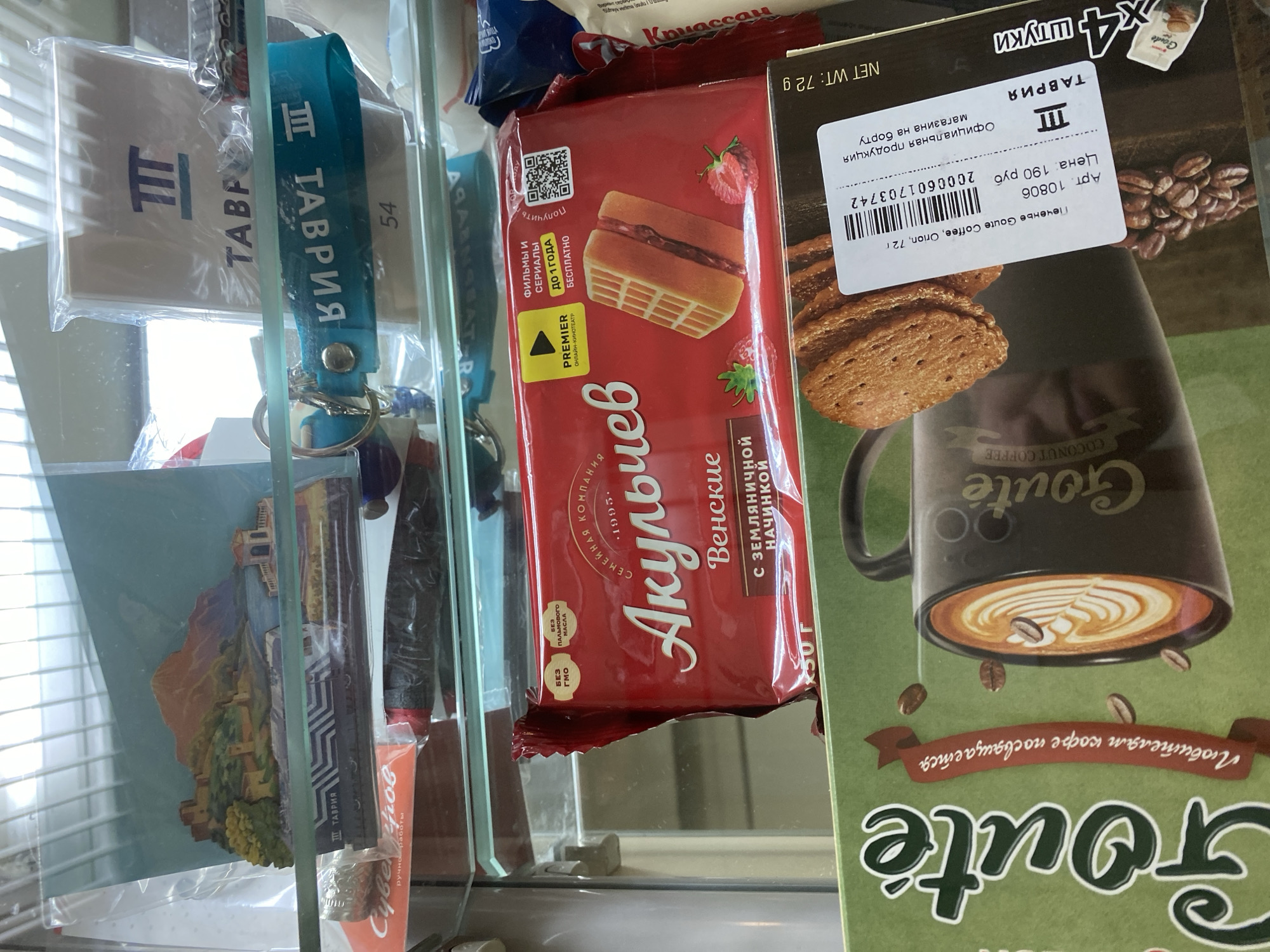 This is a close-up, eye-level photograph of items displayed on shelves, likely in a retail setting in the city of Gryazi, Russia. The foreground features a green box with the text "Goute" and an image of a black mug filled with coffee. To the right of the mug, a label with a barcode and Russian text is visible, indicating "Печенье Goute Coffee, Onion, 72 г" (Goute Coffee Onion Cookies, 72g), "Арт. 10806" (Art. 10806), "Цена. 190 руб" (Price. 190 rubles), and "Официальная продукция магазина на борту" (Official store product on board). The word "ТАВРИЯ" is also printed on the label.

Above this box, a red package of cookies labeled "Акульчев" (Akulchev) with the word "Венские" (Vienna) and "с земляничной начинкой" (with strawberry filling) is prominently displayed. Small yellow labels indicate "БЕЗ ГМО" (GMO-free) and "БЕЗ ПАЛЬМОВОГО МАСЛА" (palm oil-free). A portion of a waffle cookie with strawberry filling is depicted on the package, along with two strawberries.

Further up on the shelves, a blue keychain with white text that appears to be "ТАВРИЯ" in reverse is visible. Behind the keychain, a white box with a blue logo and the word "ТАВРИЯ" is present. To the left of the keychain, a framed souvenir depicting a landscape with a castle and water is seen. Below that, a black object with red accents, possibly a pen, is partially visible, along with a silver cylindrical object. The overall environment suggests a display of snacks and souvenirs.