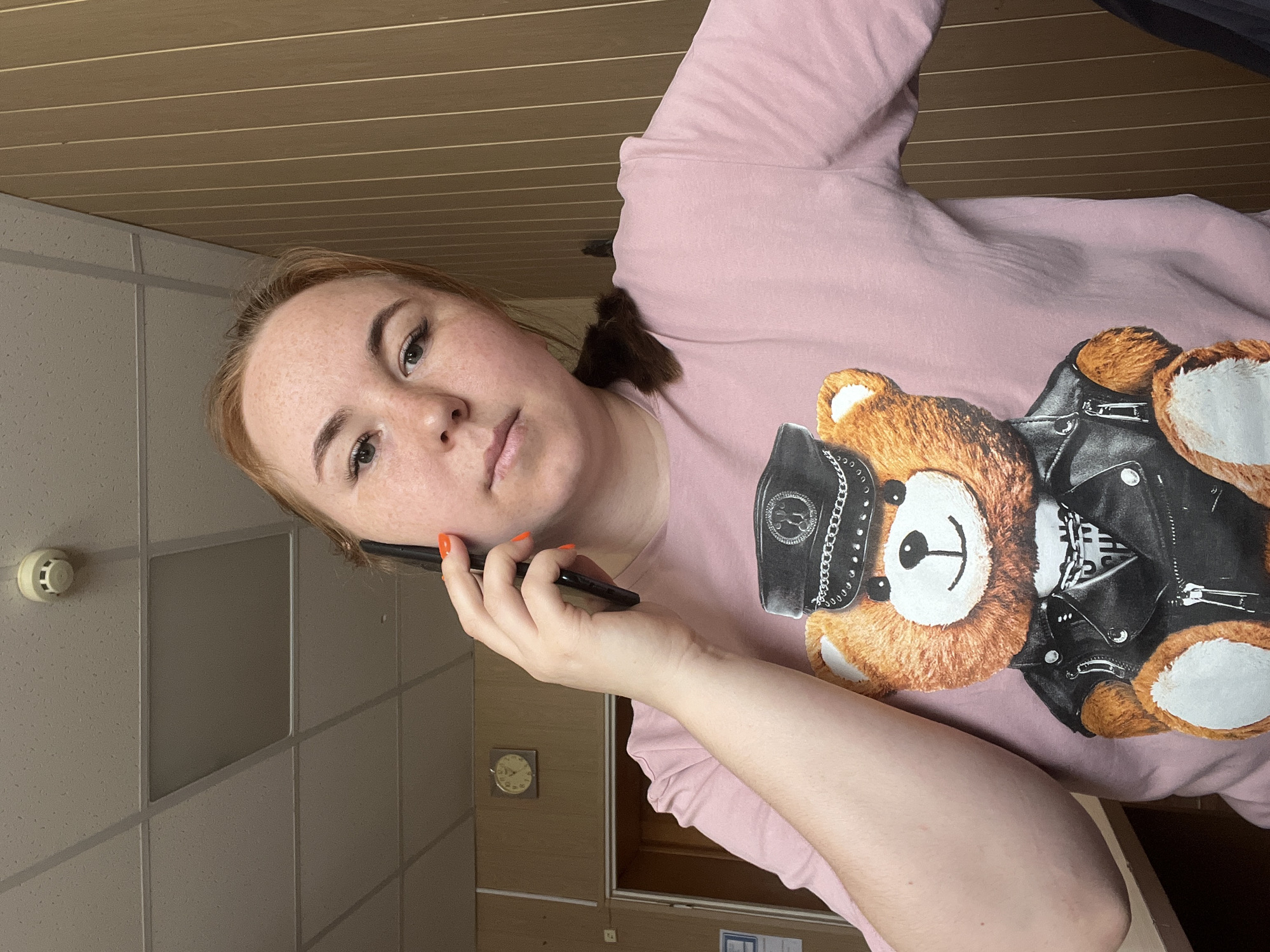 A woman is on a phone call indoors. She is fair-skinned with freckles and reddish-brown hair, wearing a pink t-shirt with a teddy bear graphic dressed in a biker jacket and hat. She is holding a black smartphone to her left ear. The background includes a drop ceiling with recessed lighting, a tiled wall, and a wooden slat ceiling. A small clock is visible on a wall, along with a fire alarm on the drop ceiling. The location may be Chaikovsky, Russia, based on the query.