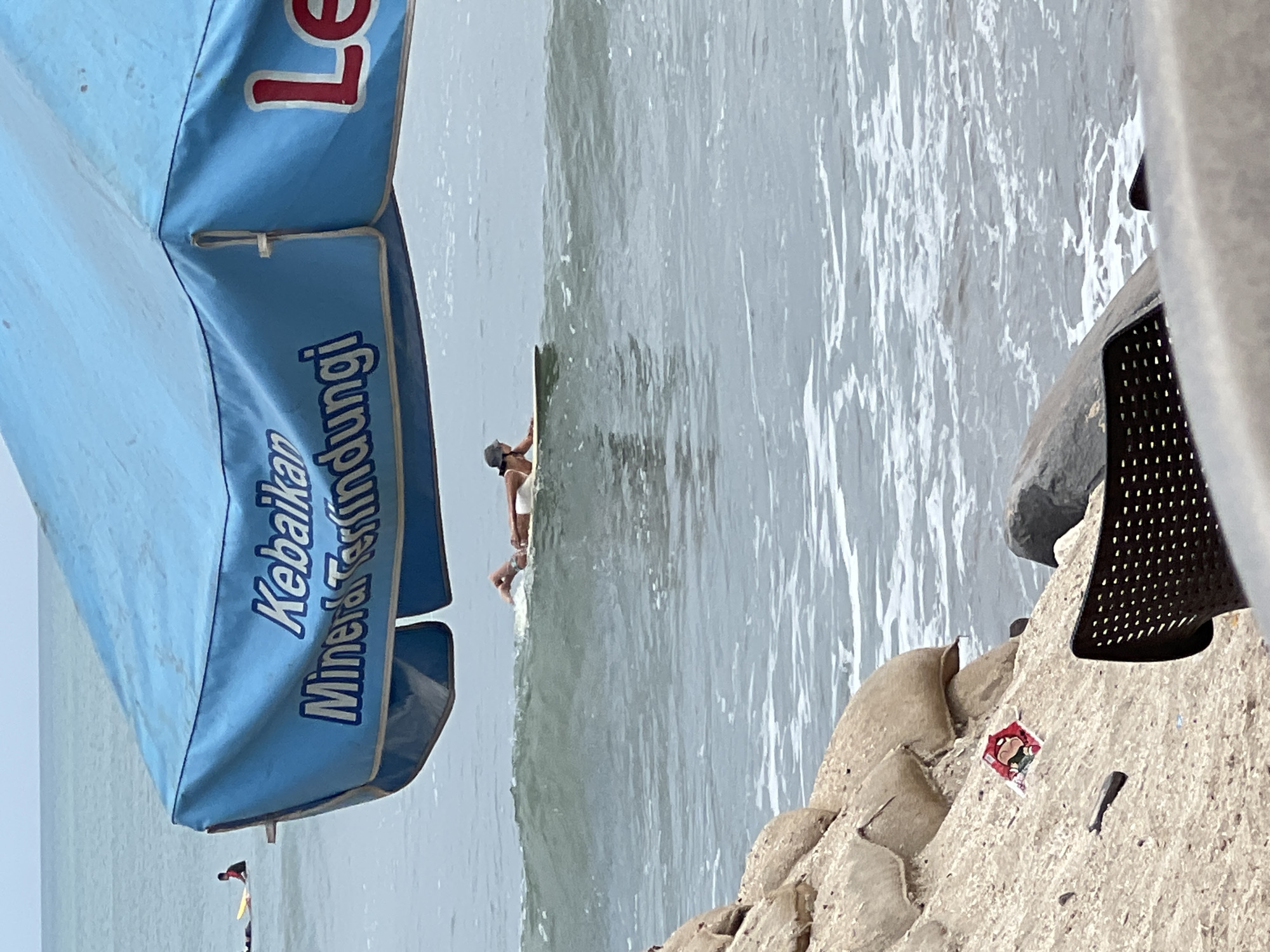 An over-the-shoulder shot reveals a serene beach scene in Kuta, Indonesia, as seen from beneath a large blue umbrella. The umbrella's fabric displays text in white, with "Kebaikan Mineral Terlindung" visible on the left side. In the distance, a surfer is captured paddling on a yellow surfboard, their body angled towards the gentle waves. Closer to the shore, another surfer, partially submerged, is seen lying on a surfboard, their legs extended towards the right. The ocean water is a calm blue, with white foam cresting small waves. The foreground is dominated by a sandy embankment, partially covered with sandbags, and a dark, perforated chair. A discarded red and white candy wrapper with a cartoon character is visible on the sand. The overall atmosphere is one of a tranquil day at the beach.