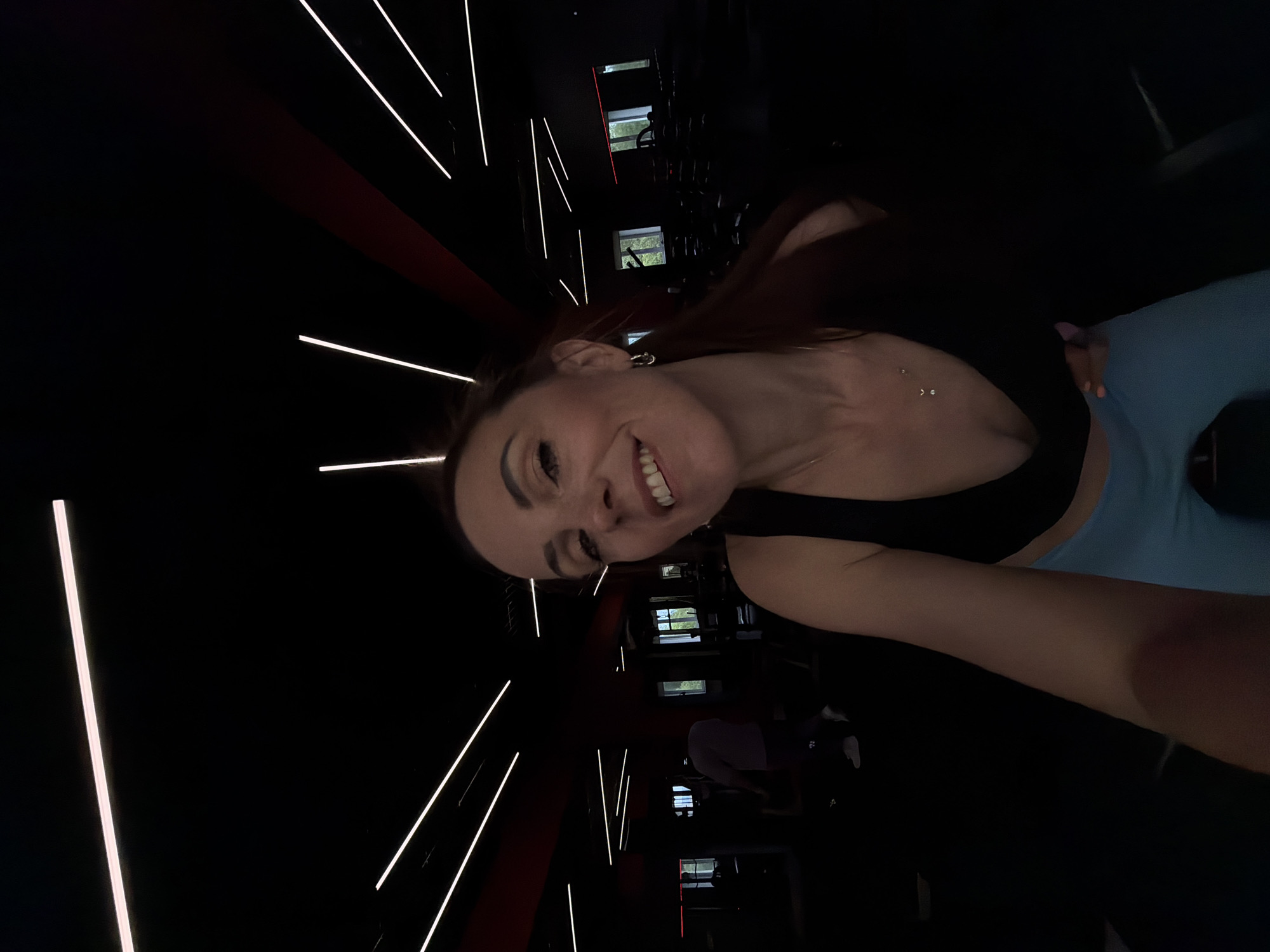 The image is a selfie taken indoors, likely in a gym. The subject is a woman with shoulder-length dark hair, wearing a black sports bra and light blue leggings. She is smiling and looking at the camera. The background shows a dimly lit gym with rows of exercise equipment and bright linear lights on the ceiling. There are a few other people visible in the background, one walking and others appearing to be using equipment. The environment suggests a modern fitness facility. The image does not contain any information that would allow for the identification of the city as Birobidzhan, Russia.
