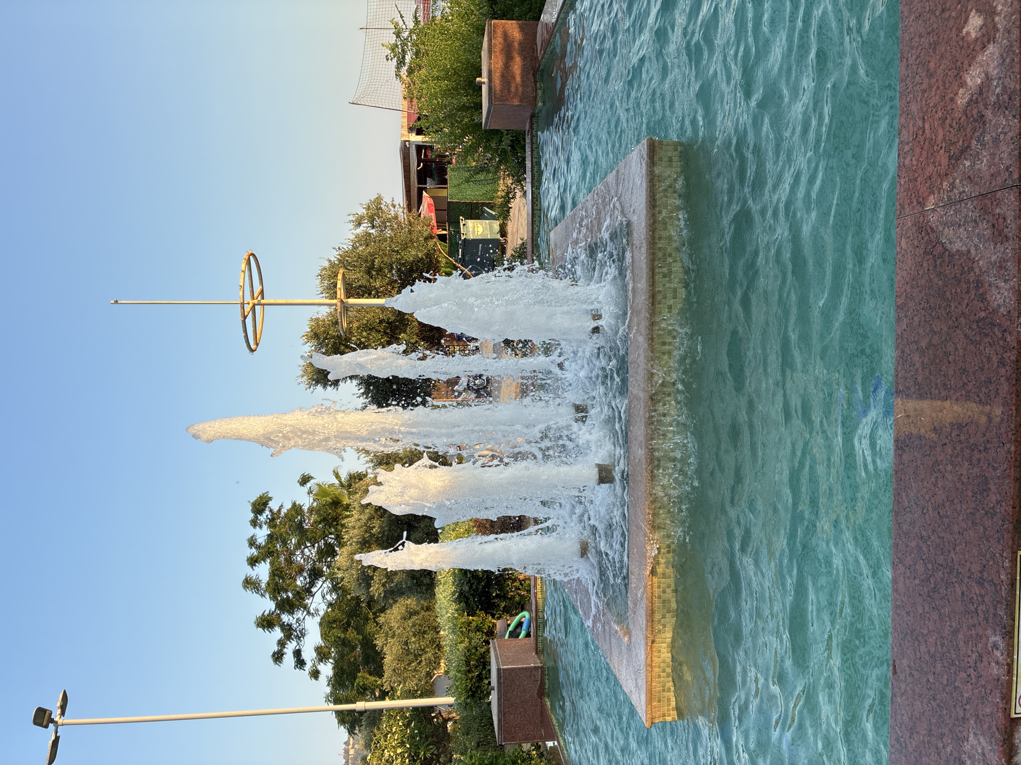 The image shows a public fountain in Alanya, Turkey. The fountain is in a pool of turquoise water, with several jets of water shooting upwards. The fountain is surrounded by landscaping, including bushes and trees. In the background, there are buildings, a street light, and a volleyball net. The sky is clear and blue, and the sun is shining.