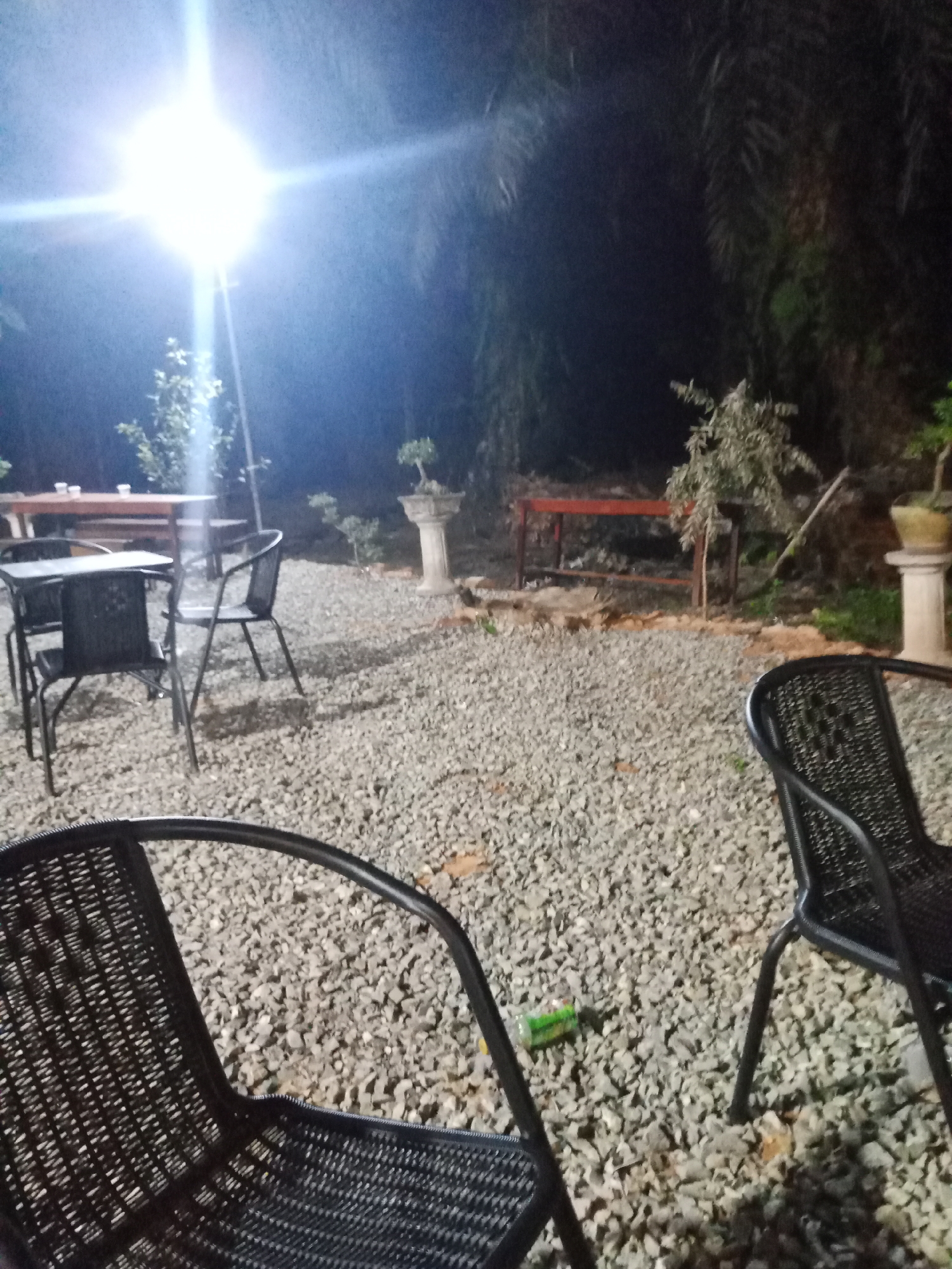 The image depicts an outdoor seating area at night, likely a cafe or restaurant, located in Mekar Jaya, Indonesia. A bright overhead light illuminates the scene, casting shadows on the gravel ground. Several black, woven chairs and tables are arranged outdoors. In the background, a long wooden table with cups on it is visible, along with a red bench. Decorative elements such as a potted plant on a pedestal and a bonsai tree are also present. The overall ambiance suggests a tranquil outdoor dining or relaxation space.