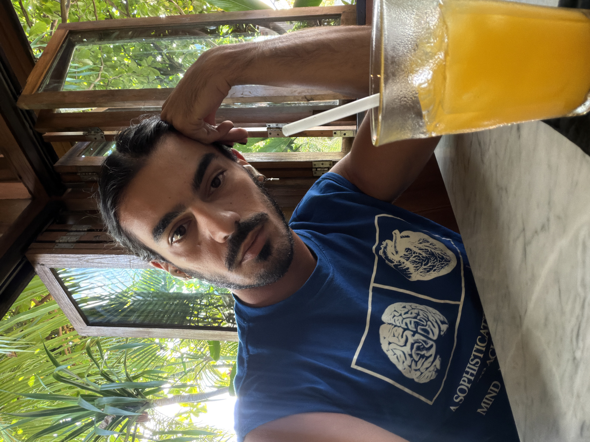 In North Kuta, Indonesia, a man with dark hair and a beard is seated at a table, resting his head on his hand and looking at the camera. He is wearing a blue t-shirt with an image of a brain and a heart, and the text "A SOPHISTICAT MIND ... ACT". In front of him is a glass of orange juice with ice and a straw. The background shows lush green foliage and wooden window frames, suggesting an outdoor or semi-outdoor dining area. The man's left arm is bent and his hand is supporting his head. He is also wearing a small cross earring.