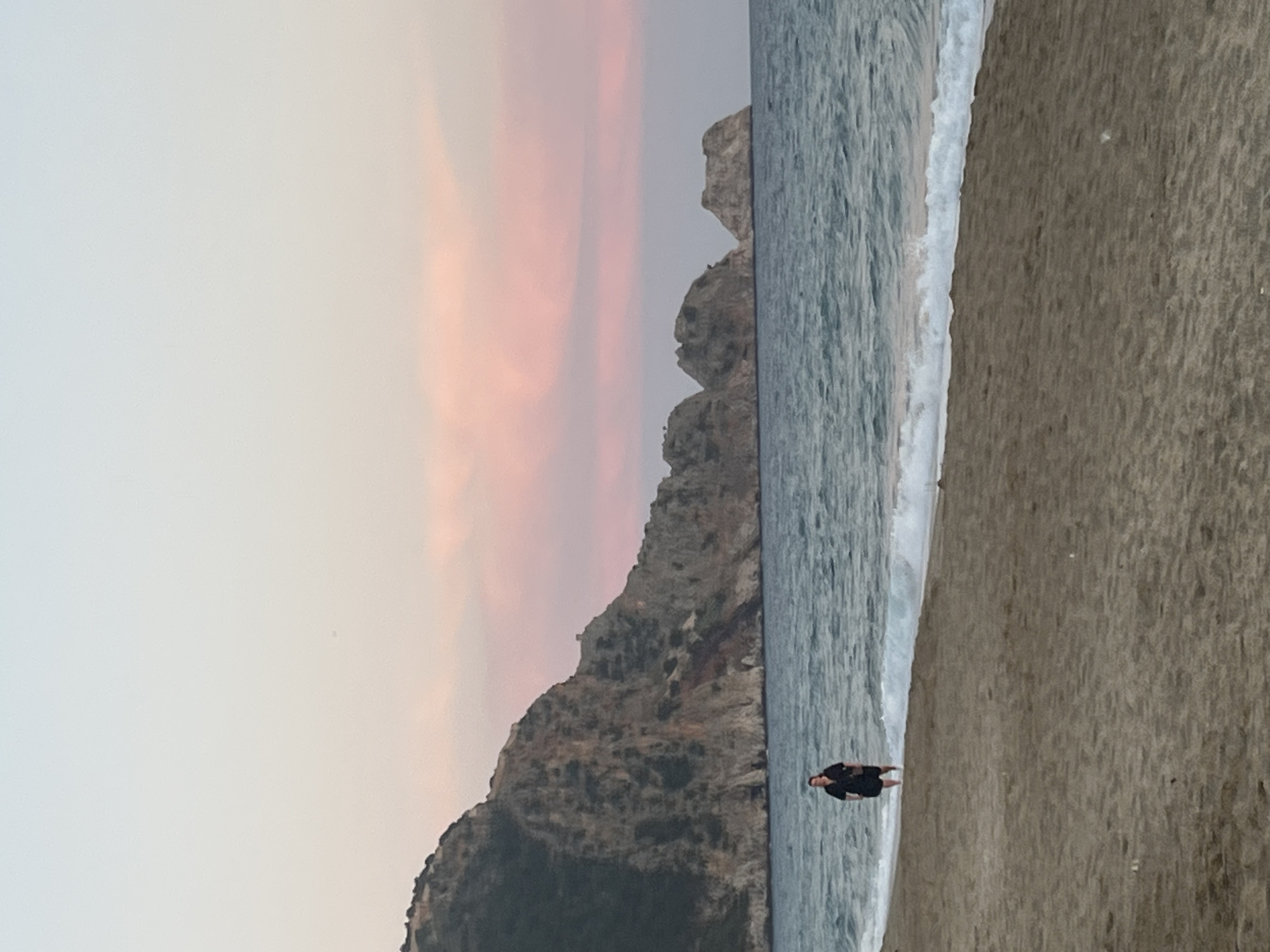 A beach scene, likely near Alanya, Turkey, shows a figure standing on the sand where it meets the water. The beach is sandy, and the water is light gray, possibly due to the light. A mountain or large rock formation is visible along the water's edge in the middle distance. The sky is a mix of light gray and soft pink hues, suggesting sunset or sunrise. The image is rotated 90 degrees.