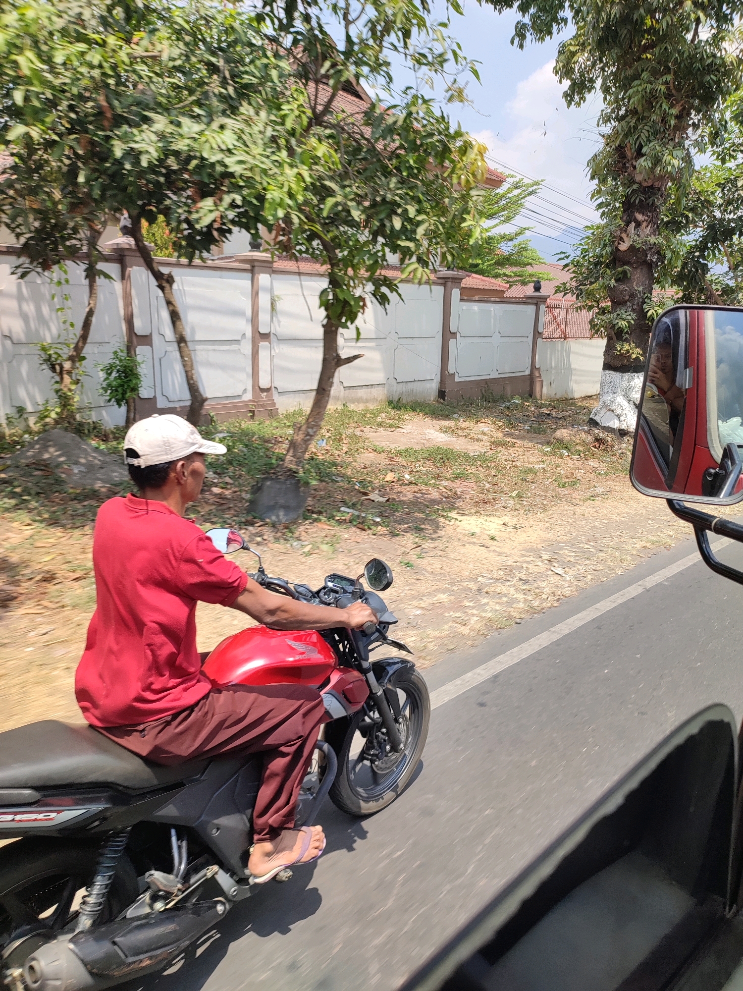 In Salaman, Indonesia, a man wearing a red shirt and brown pants rides a red motorcycle on a paved road. He is wearing a white baseball cap and light-colored flip-flops. The motorcycle has a black seat and black accents. The background shows a white fence with decorative posts, trees with green foliage, and a residential building with a terracotta roof. The sky is partly cloudy. The image is taken from the perspective of another vehicle, as evidenced by the reflection of the photographer in the motorcycle's side mirror.