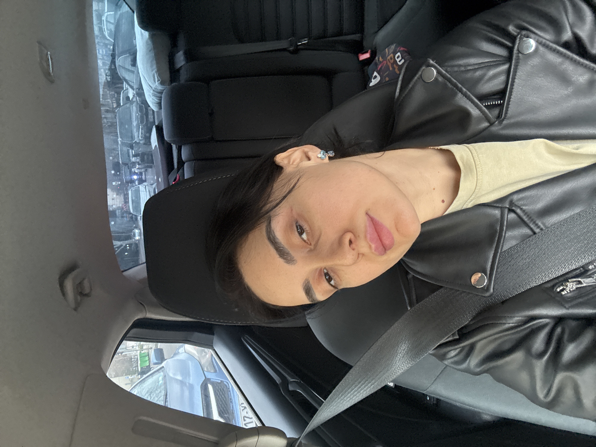 A forensic examination of the image reveals a female individual occupying the front passenger seat of a vehicle, taking a selfie. She has dark hair, dark eyebrows, and is wearing a black faux-leather jacket over a light-colored t-shirt. A grey seatbelt crosses her chest. A blue and silver floral or butterfly-shaped earring is visible on her right ear. Her expression is neutral with slightly parted lips.
The interior of the vehicle features dark upholstery. The headrest of the front passenger seat is visible, and the rear seats appear dark, with a white pillow and a patterned object (possibly a bag or fabric) placed on them.
Through the front passenger window, a portion of a blue Subaru Forester (or similar SUV) is visible, displaying a license plate with the partial sequence "17-V". Green urban-style trash bins are visible further down the street on the left. Through the rear window, a busy urban street with moderate traffic is observed, indicating a congested area. Multiple vehicles, including a dark SUV (possibly a BMW X-series), are visible. Trees line the street, and multi-story buildings are faintly discernible in the background.
Based on the prompt and the partial license plate sequence visible on the adjacent vehicle, the scene is situated on an urban street within the city of Tbilisi, Georgia, specifically amidst traffic. No further specific street or landmark identifiers are discernible from the visual evidence.