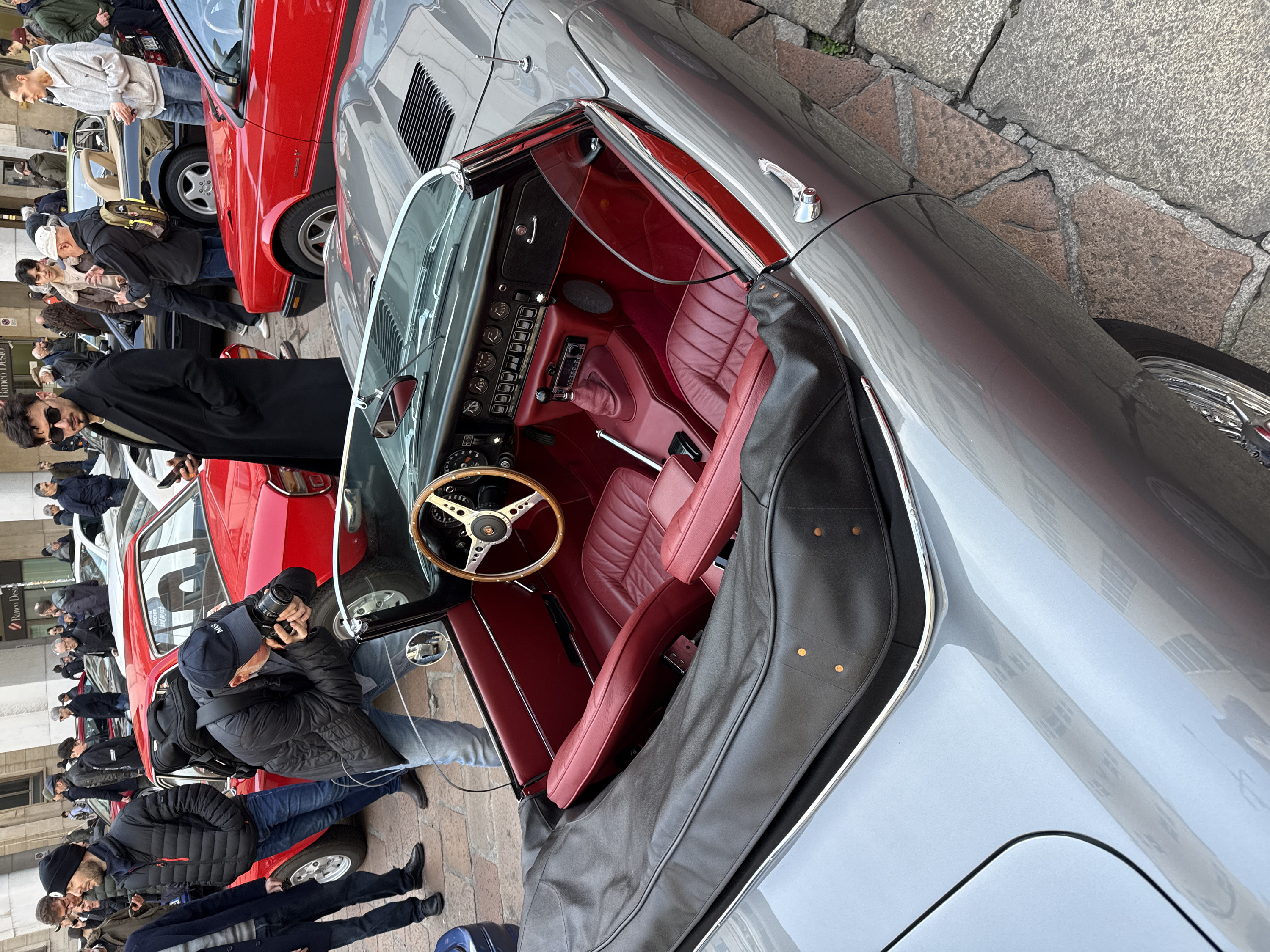 The scene is set on a cobblestone-paved public street or square in Milan, Italy, featuring a display of classic automobiles. In the foreground, a grey classic convertible sports car, identifiable as a Jaguar E-Type, is prominently featured. Its soft-top is partially covered by a dark grey tonneau cover, and the interior boasts rich red leather seats, a matching lower dashboard, and a polished wooden steering wheel. The dashboard is equipped with multiple analog gauges, toggle switches, and an aftermarket digital radio. The driver's door is ajar, revealing the red interior.
Approximately fifteen to twenty individuals, mostly adult males, are distributed in the mid-ground and background, observing the vehicles. A man wearing a dark baseball cap and jacket is actively photographing the grey convertible with a professional camera, adopting a crouched posture. Nearby, a man with dark hair and sunglasses, dressed in a black long coat, stands upright while holding a smartphone. Other individuals are standing, observing, or walking amidst a collection of classic cars, including a red coupe partially visible behind the photographer, another red classic car to the right, a white convertible, and a black convertible with a beige interior further in the background. The background features multi-story urban buildings, one of which displays a "Banco Desio" sign, and prominent architectural columns. The ground is paved with irregular grey and reddish-brown cobblestones.