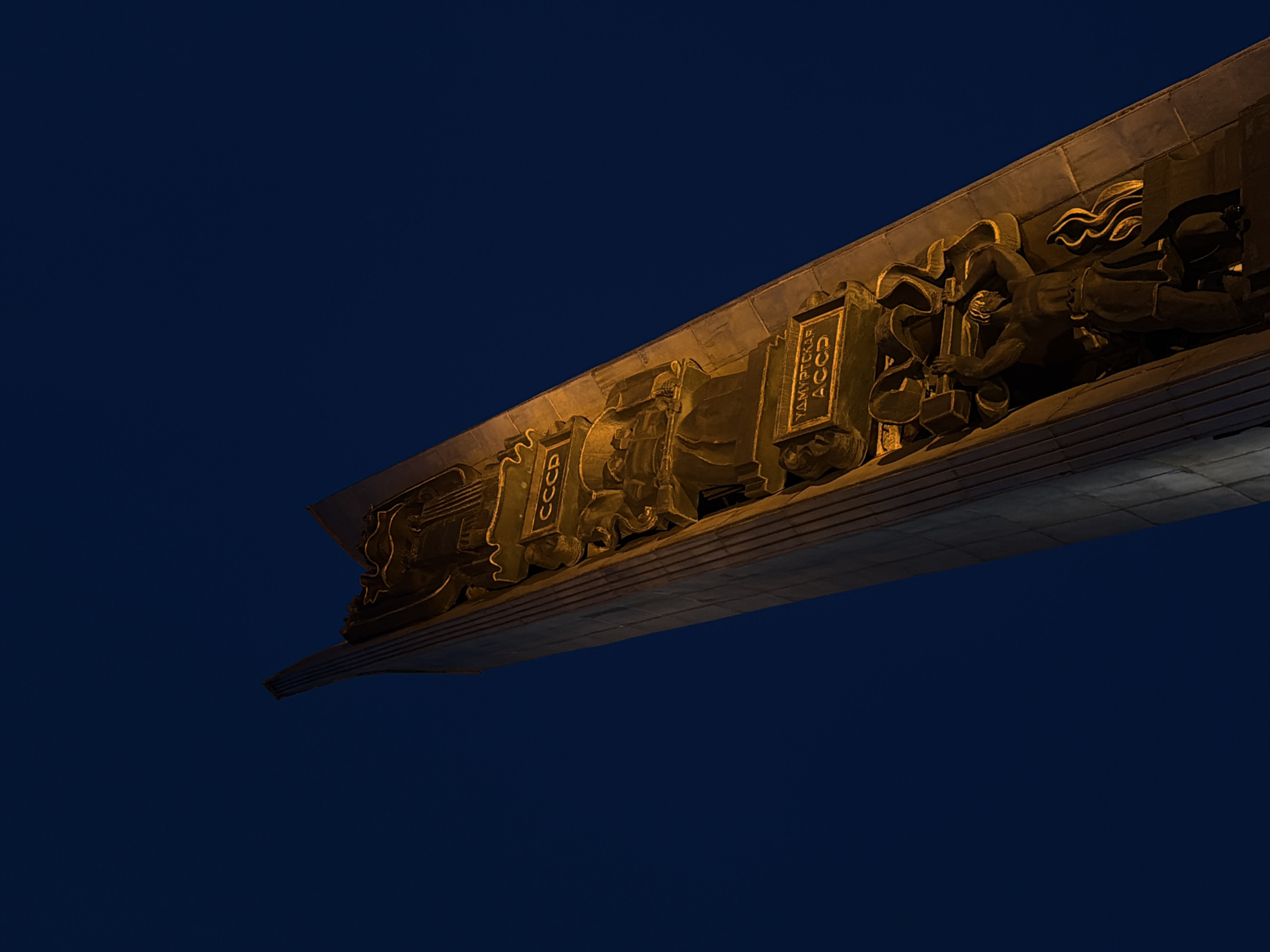 The image shows a close-up of the monument to the friendship of peoples in Izhevsk, Russia, at night. The architectural structure features golden bas-relief sculptures against a dark blue sky. One of the reliefs contains the Russian abbreviation "CCCP." The monument is illuminated with artificial light.