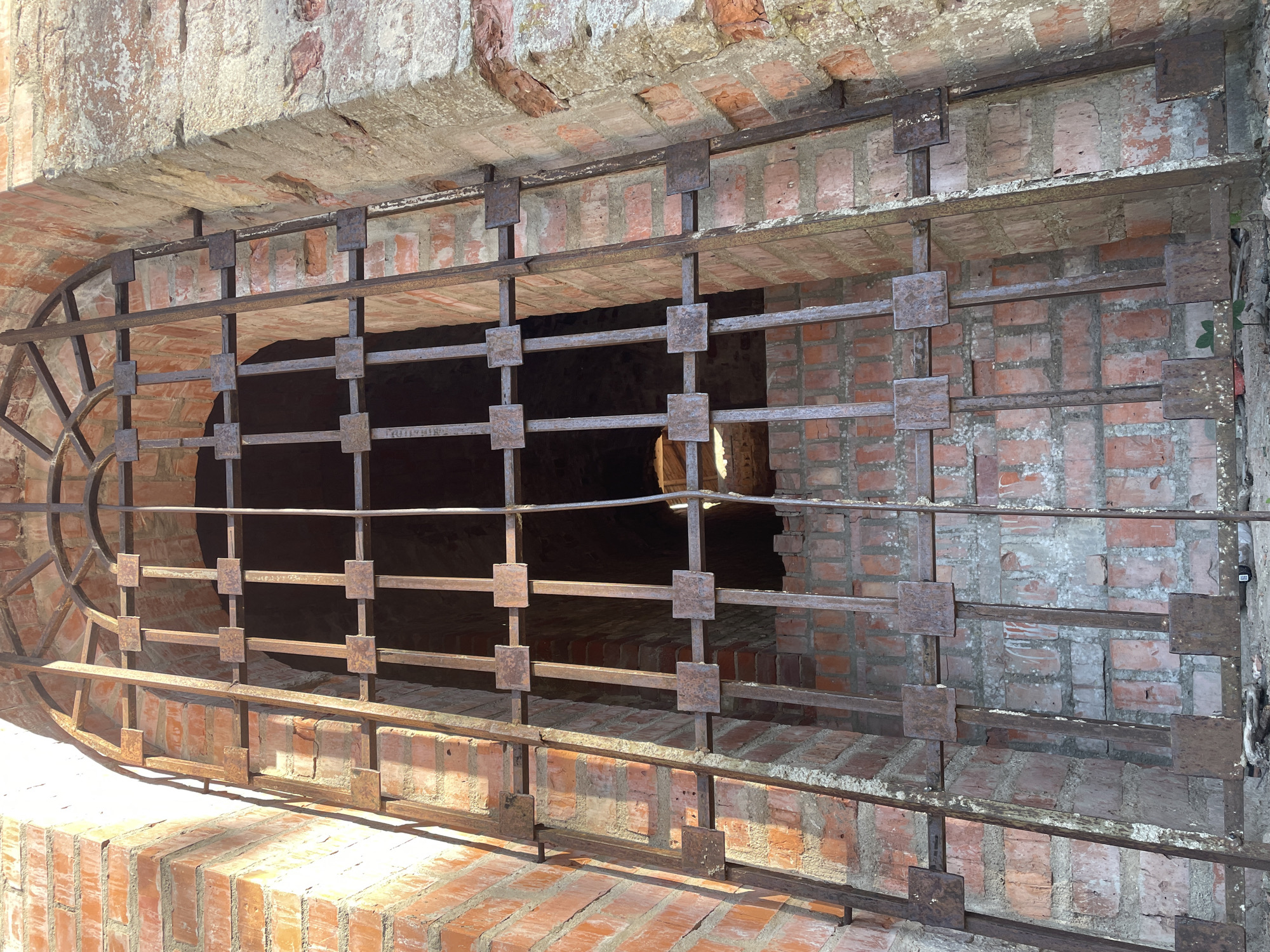 The image shows a close-up, low-angle view of a large, arched, wrought-iron gate set into a brick structure. The gate is made of thick, vertical bars connected by horizontal crossbars, with square plates at the intersection points. The metal is heavily rusted. The brickwork behind and around the gate is rough and appears aged, with some areas of crumbling mortar. Sunlight streams in from the top right, casting strong shadows across the gate and the bricks. The scene is devoid of people or any signs of recent activity. The overall impression is one of abandonment and decay. The location is identified as Sof'ino, Russia.