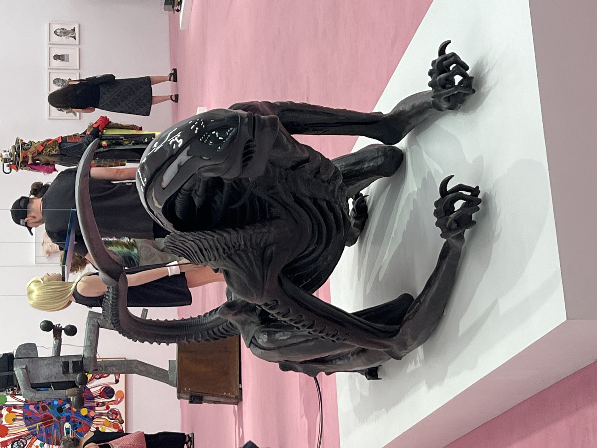 This image captures a sculpture of a creature, resembling a Xenomorph from the "Alien" franchise, displayed on a white pedestal. The sculpture is dark, glossy, and exhibits intricate details of its skeletal structure and clawed limbs. It is positioned on a pink carpeted floor, suggesting an exhibition or gallery setting. In the background, other artworks and people are visible. To the left, a colorful, abstract sculpture stands near a person in a black dress. Further back, a group of people are observing artworks on a white wall, including three framed pieces. The scene appears to be indoors, with bright lighting. The presence of art, people in casual attire, and the exhibition-like arrangement strongly suggest this scene is taking place at an art fair or gallery, potentially within New York City, United States of America, known for its vibrant art scene and numerous exhibition spaces.