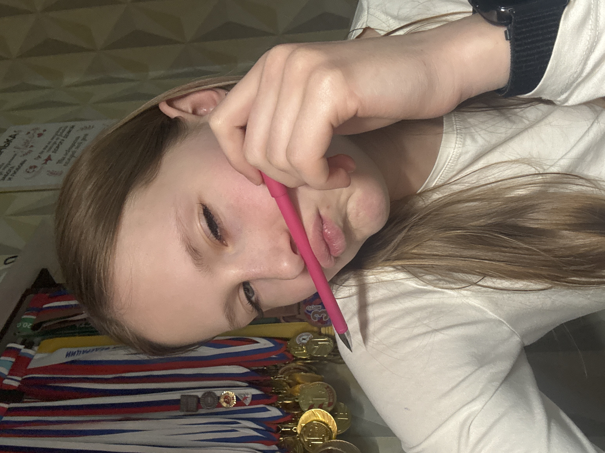 An indoor, tilted photograph depicts a young female, likely a teenager, with light brown hair, wearing a white long-sleeved shirt and a black smartwatch on her left wrist. She holds a pink pen between her upper lip and nose, looking downwards with a pensive or bored expression.
The scene appears to be a personal space, possibly a room in Chaikovsky, Russia, given the context and visible cues. The foreground and mid-ground are dominated by numerous gold medals with red, white, and blue ribbons, strongly resembling the colors of the Russian flag, suggesting significant achievements, likely in sports or academics. One medal clearly shows the Cyrillic word "MECTO" (Place). Below the medals, a striped fabric with similar red, white, and blue colors is visible. To the upper left, partially obscured by the person's head, are papers or certificates, one of which partially displays the Cyrillic word "БЛАГОДАРСТВЕННОЕ" (Gratitude/Thank You).
The background features a geometrically patterned wall. The lighting is artificial, suggesting the image was taken indoors, possibly in the evening or at night. The activity centers around the person's pose with the pen, perhaps indicating a moment of thought or idleness.