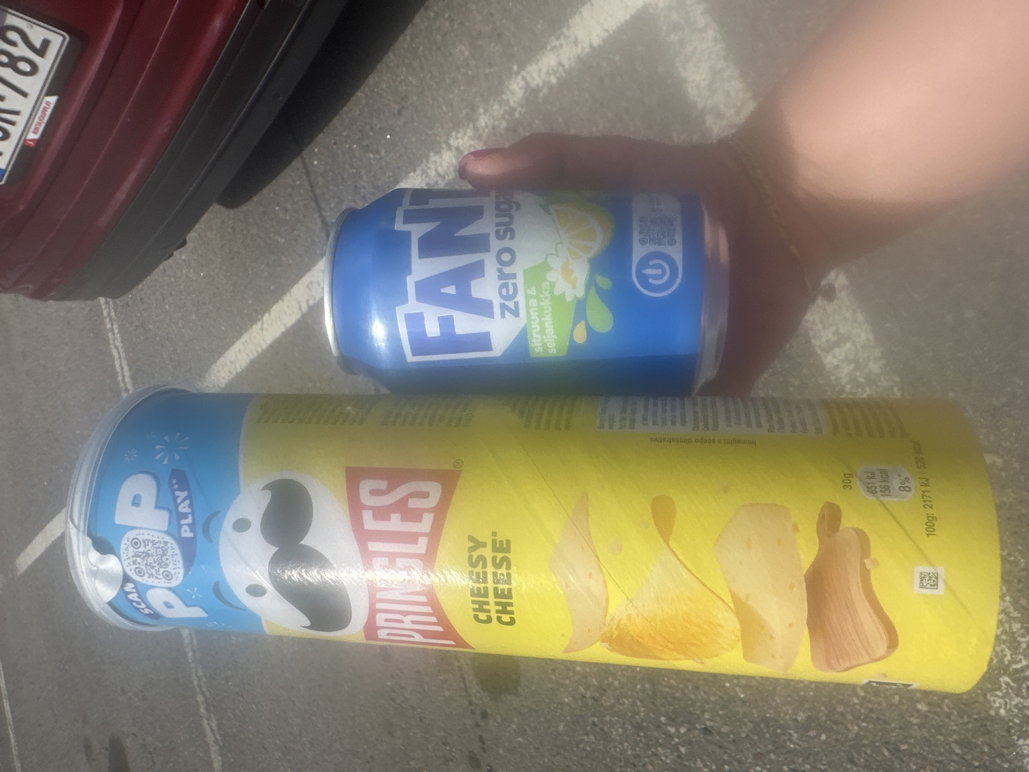 This is a close-up, slightly blurry, outdoor photograph taken from a low angle. The primary subjects are two food items: a can of Fanta Zero Sugar and a Pringles can. The Fanta can is blue and features the Fanta logo, along with text indicating "zero sugar" and a lemon and flower graphic. The Pringles can is yellow and blue, with the Pringles logo prominently displayed. The text on the Pringles can identifies the flavor as "CHEESY CHEESE" and also includes "POP PLAY" and a QR code. Nutritional information and the word "SCAN" are also visible on the can.
In the background, a red car's bumper and license plate are visible, suggesting the photo was taken in a parking lot or near a vehicle. The ground appears to be asphalt with white painted lines, reinforcing the parking lot setting. A person's hand, with a visible fingernail, is seen holding the Fanta can. The lighting suggests it is daytime, with some harsh glare on the Fanta can. The overall scene is casual and seems to capture a moment of convenience or snacking.